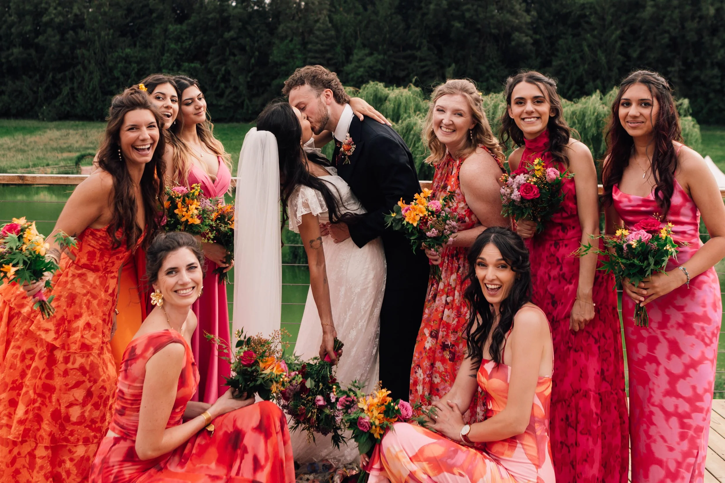 A group of people, including a bride and groom, pose for a photo at an outdoor wedding with vibrant dresses for the women, flowers, and greenery in the background.
