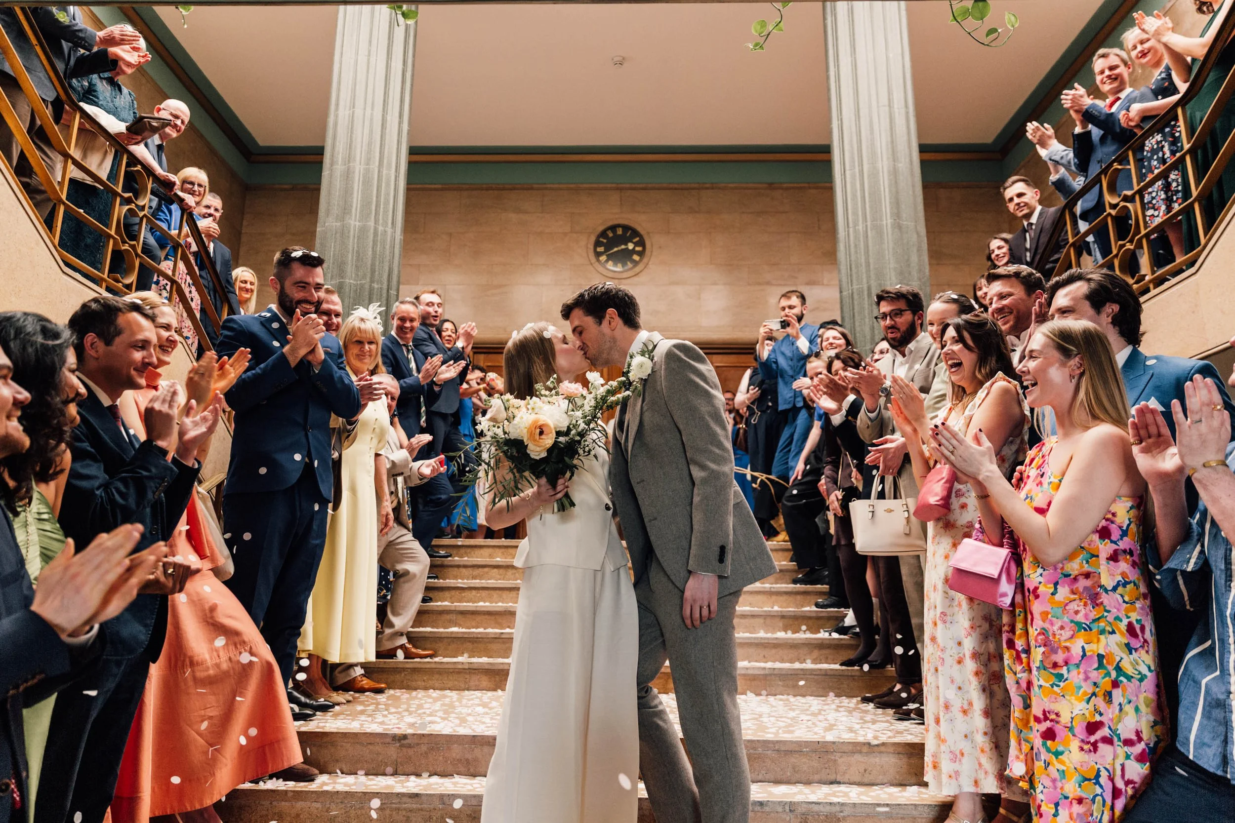 A wedding is taking place on a staircase with the bride and groom kissing, surrounded by smiling guests clapping and celebrating.