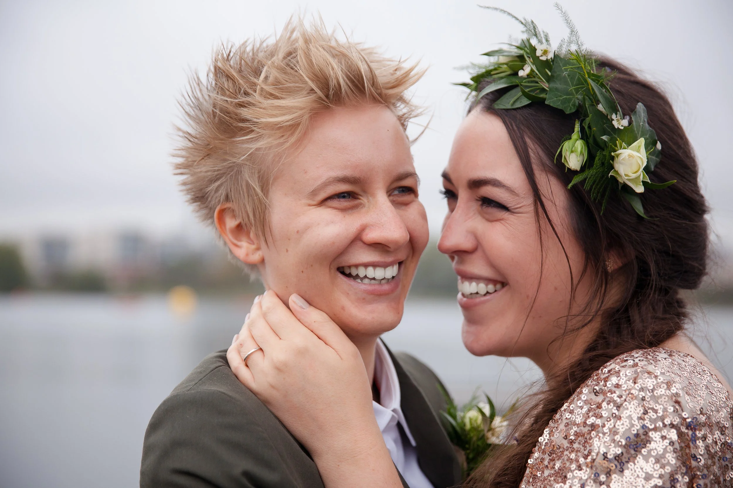 Two women smiling and embracing each other outdoors, one wearing a flower crown, with a body of water and city skyline in the background.