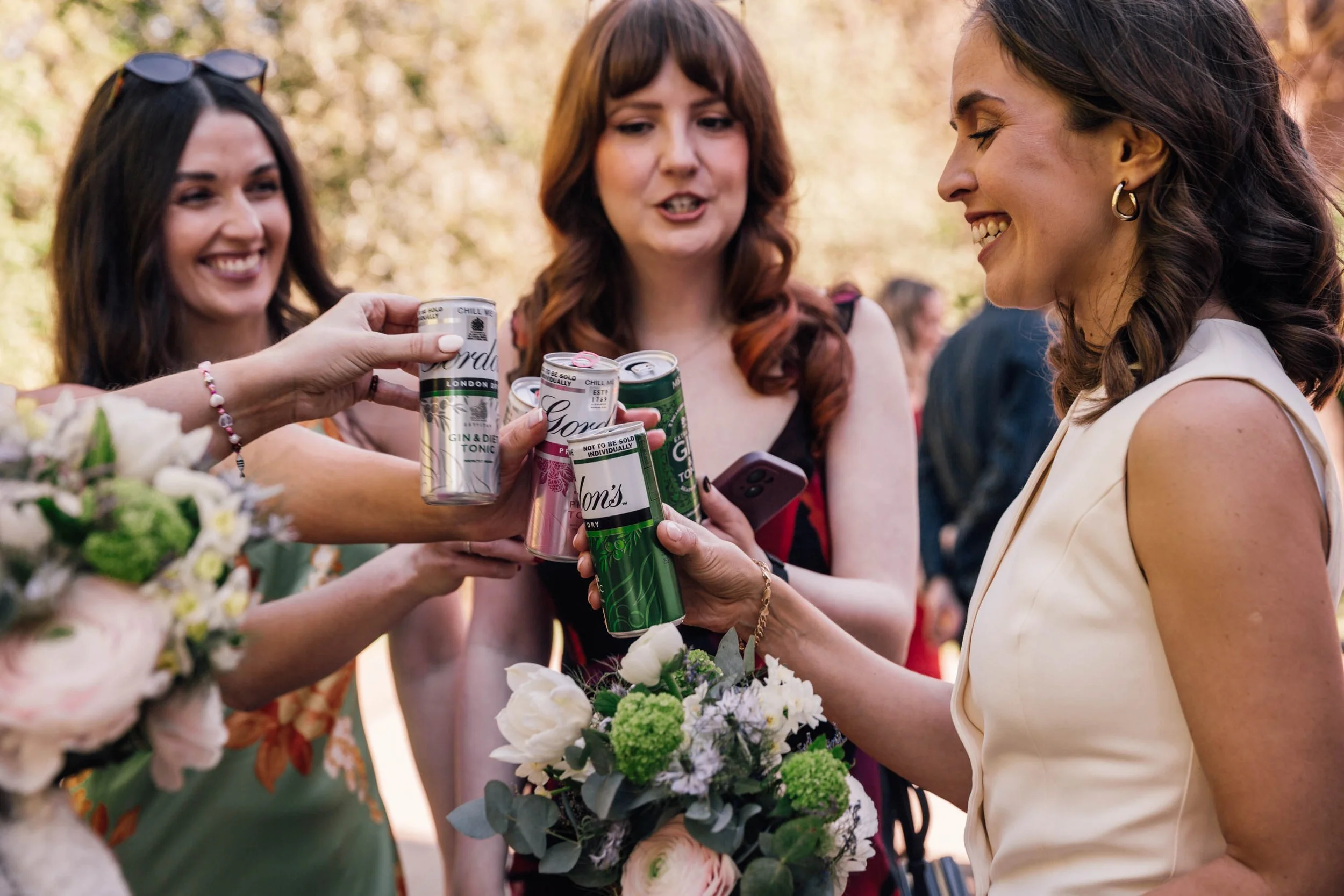 Women gathered outdoors during daytime, exchanging drinks from cans and holding a bouquet of flowers, with trees in the background.