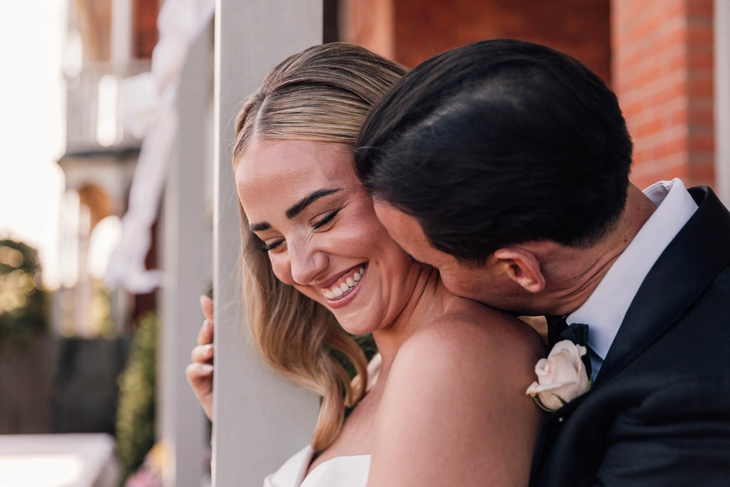 A close-up of a couple, smiling with eyes closed, leaning their foreheads together during their wedding, with a church in the background.