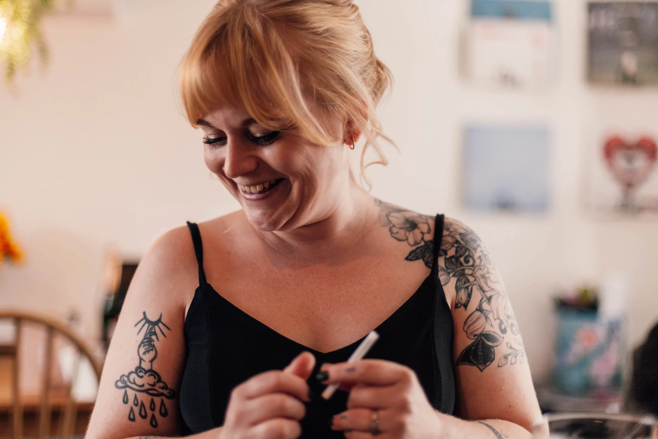 A smiling woman with tattoos on her arms and chest, wearing a black top, looking down at her phone.