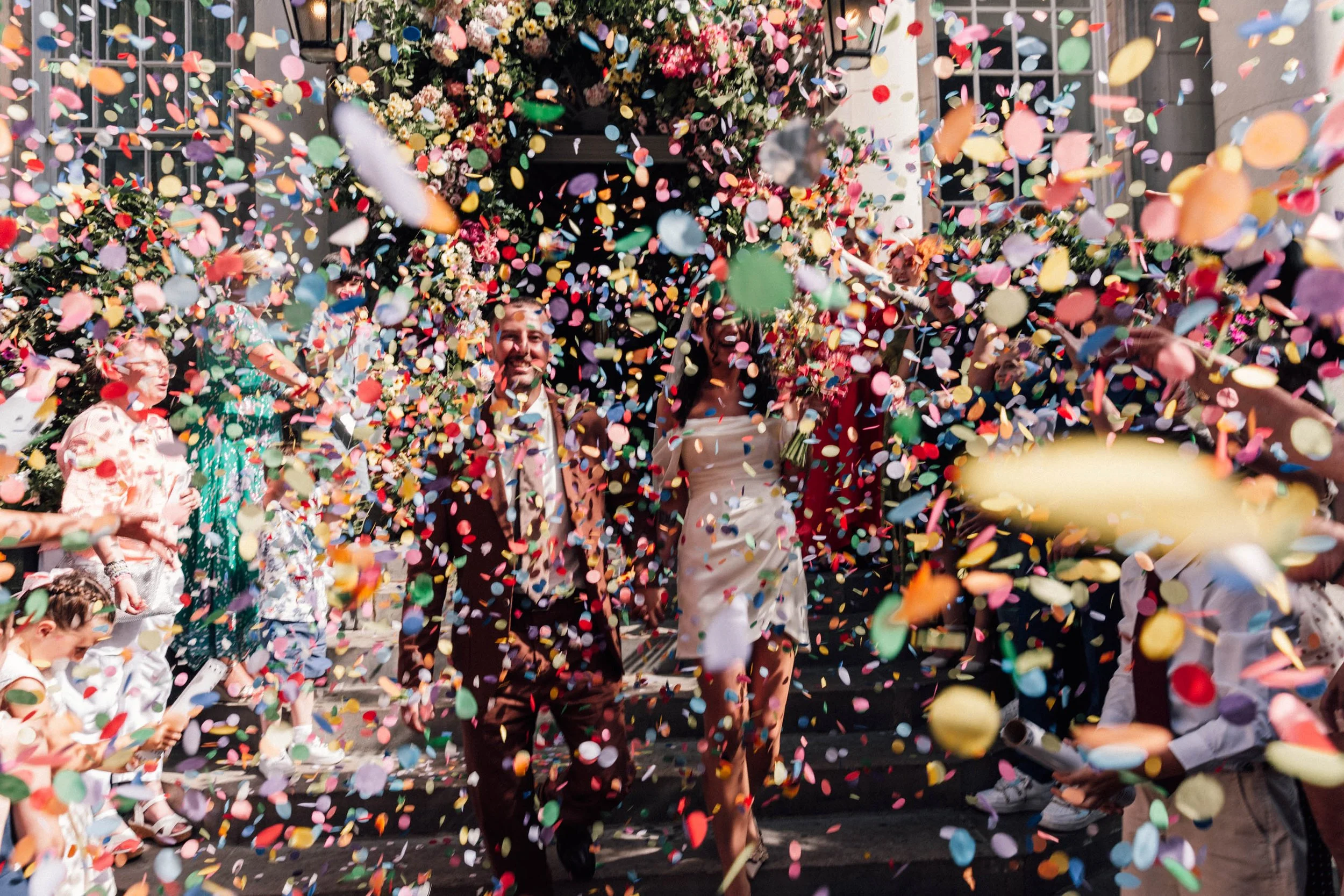 People celebrating at a wedding, throwing colorful confetti in the air with a floral backdrop and buildings in the background.