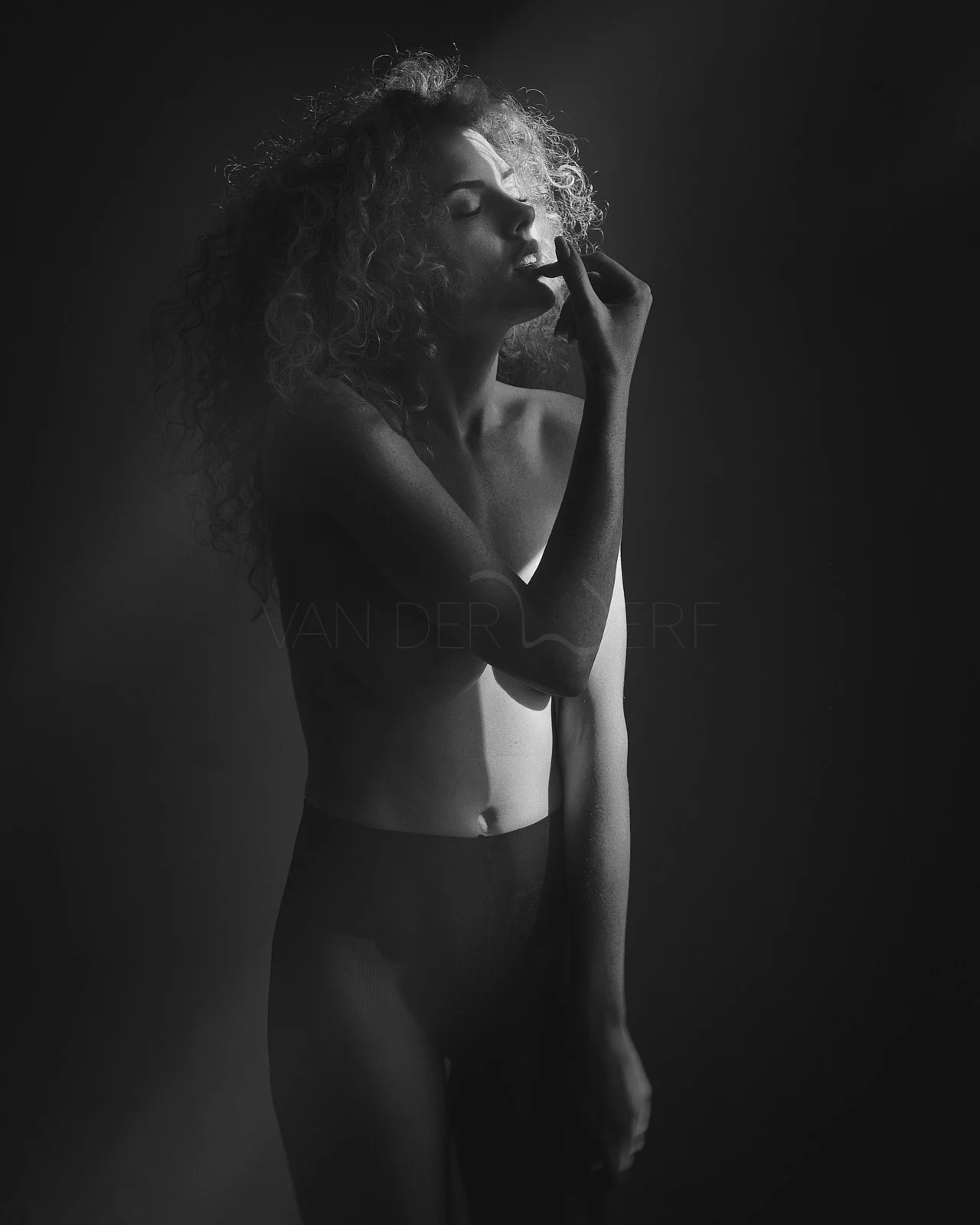 Black and white photo of a woman with curly hair, standing topless with her eyes closed and one hand touching her lips, in a moody lighting setting.