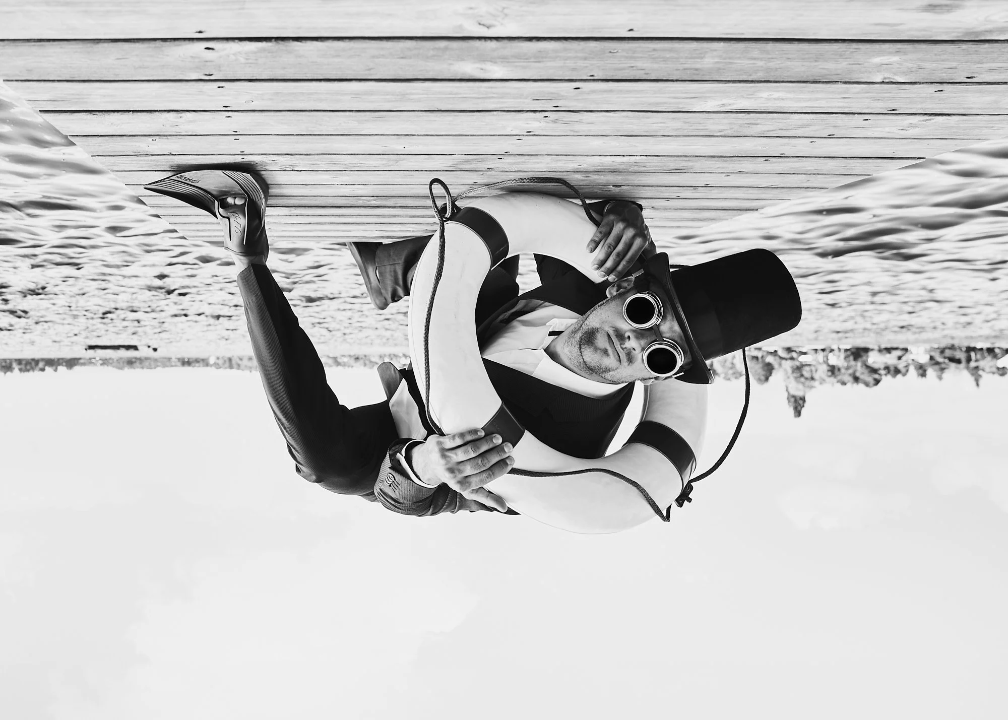 Man wearing a top hat, goggles, and a tuxedo, holding a life preserver, leaning over a wooden dock with a body of water and a bridge in the background, in black and white.