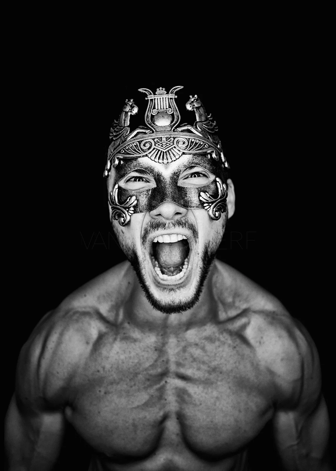 Black and white photo of a muscular man wearing an ornate, decorative mask with a fierce expression, mouth open as if shouting, against a black background.