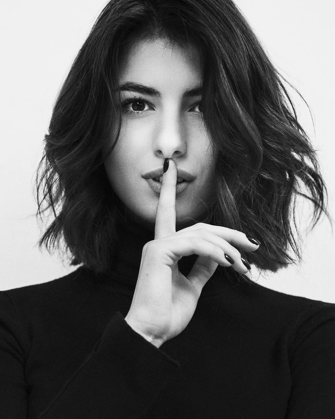 Black and white portrait of a woman with short wavy hair, looking directly at the camera, holding her index finger to her lips in a shushing gesture.