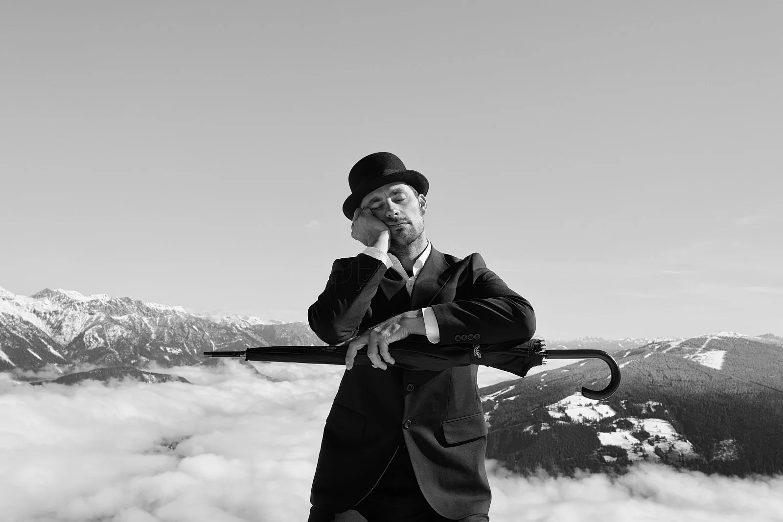 Black and white photo of a man in a suit and bowler hat, resting his head on his hand with eyes closed, holding a closed umbrella with the other hand, standing in front of mountains and clouds.