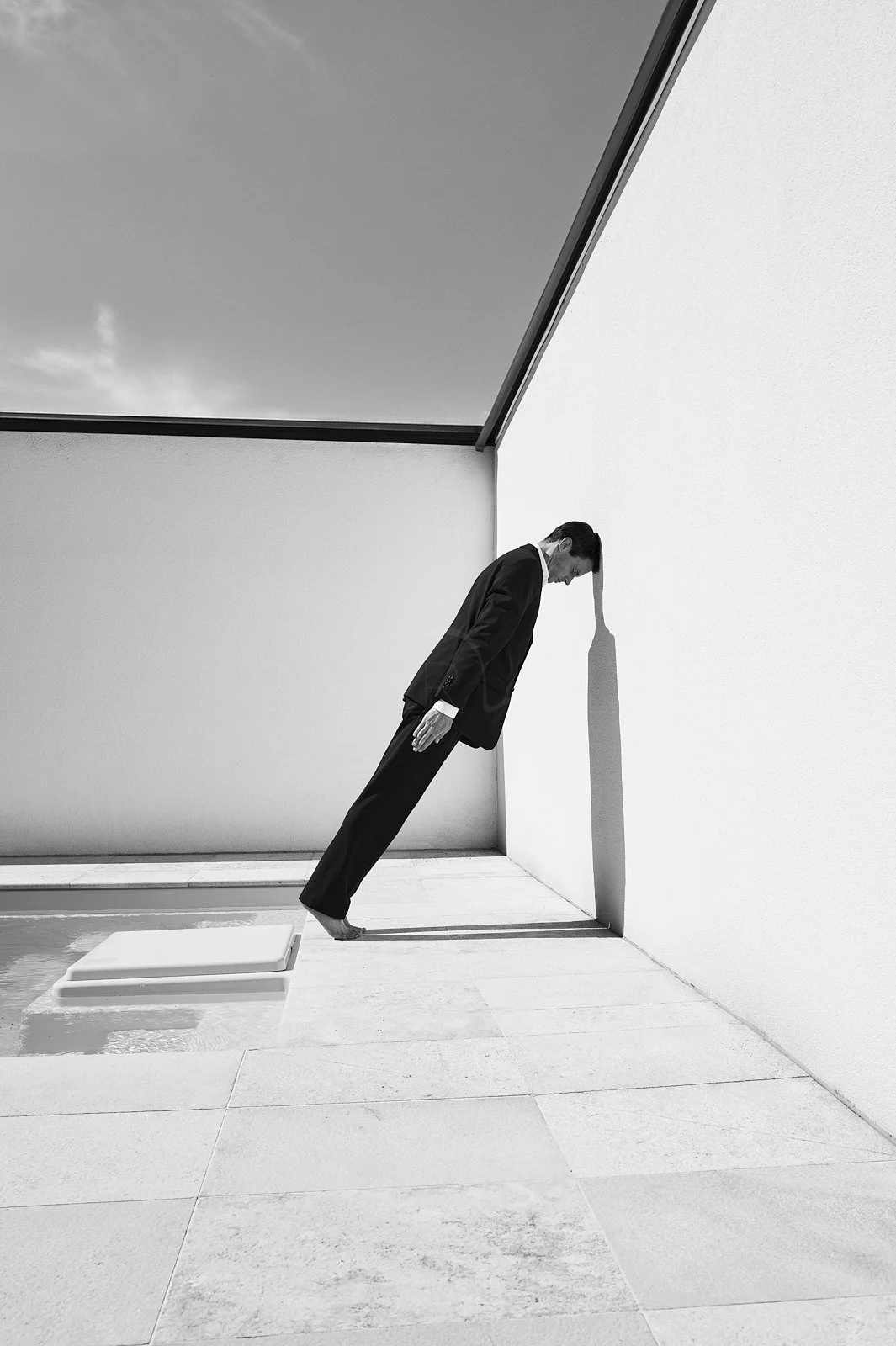A man in a suit leaning against a wall, with his head and shadow extending downward in a surreal, tilted scene.