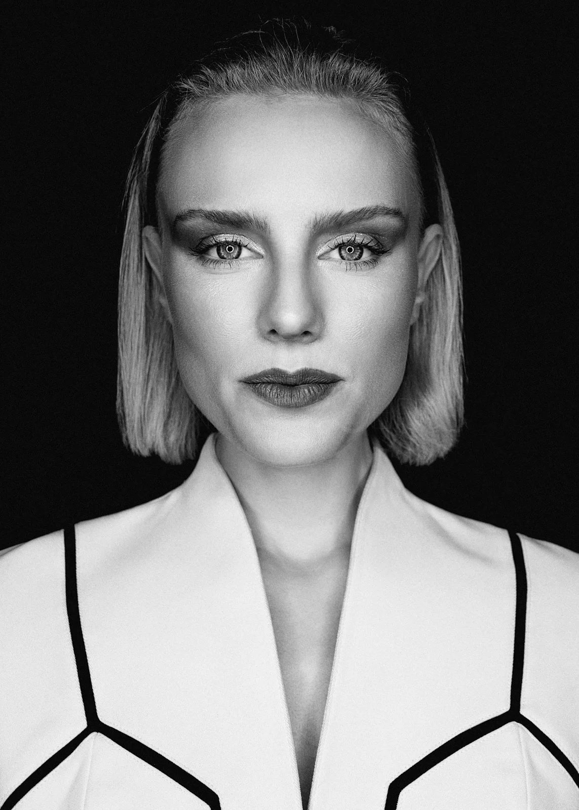 Black and white portrait of a woman with short blonde hair, wearing a geometric patterned top, looking directly at the camera against a dark background.