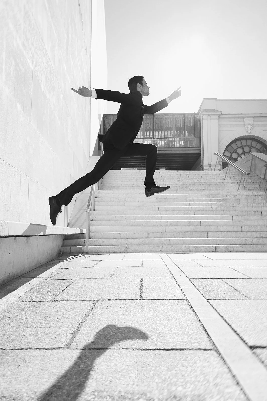 A man in a business suit appears to be leaping up a staircase outdoors, with one leg on the ground and the other mid-air, with his arms extended outward. The photo is in black and white, with strong sunlight casting shadows.