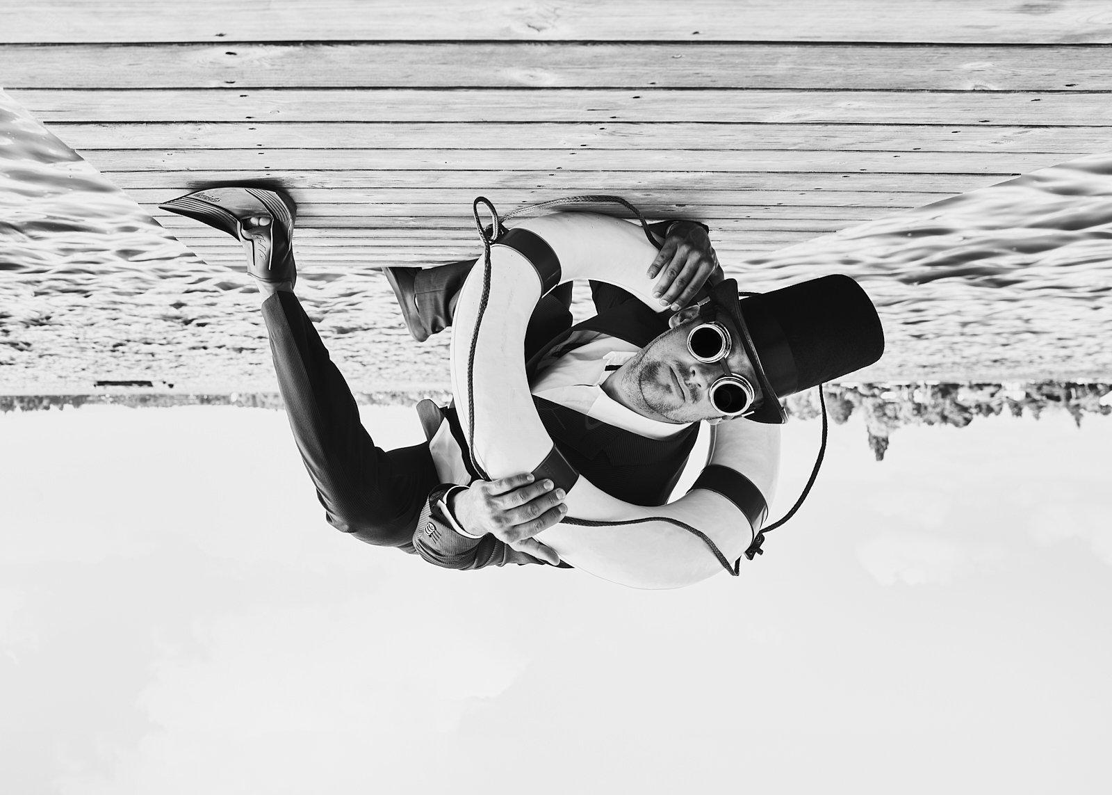 Man in steampunk costume with top hat and round sunglasses sitting on a beach, holding a life-saving buoy.