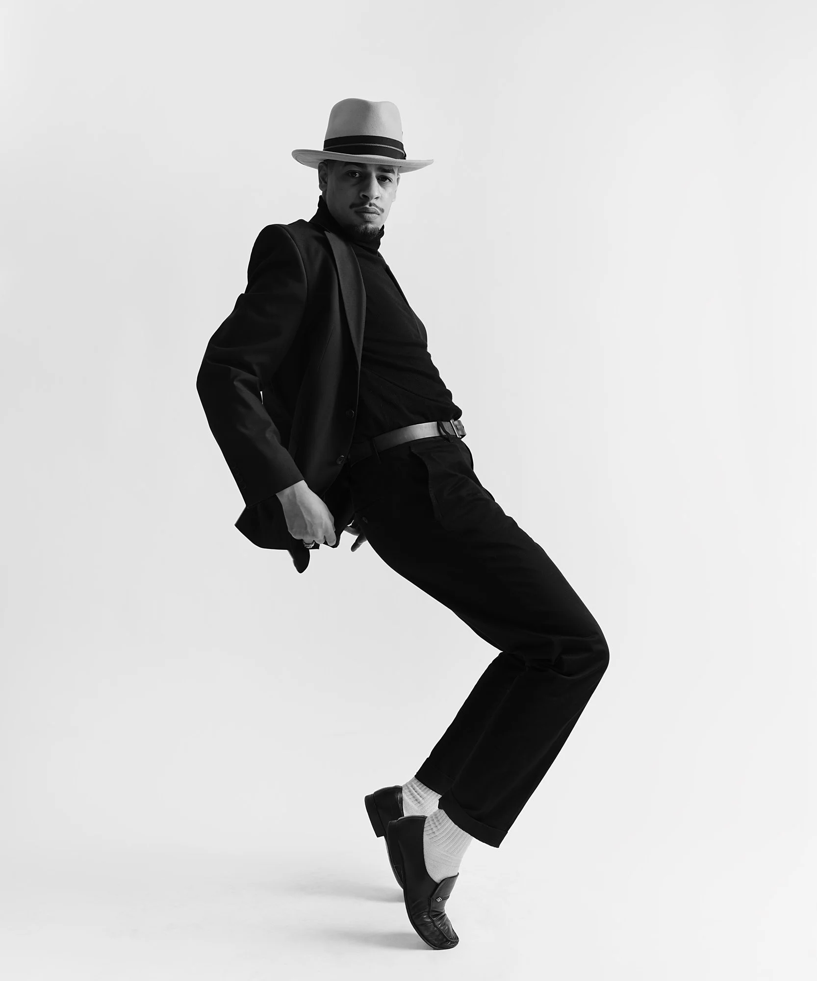A man wearing a fedora hat, dark blazer, tucked-in shirt, dark pants, socks, and loafers, posed against a plain background.