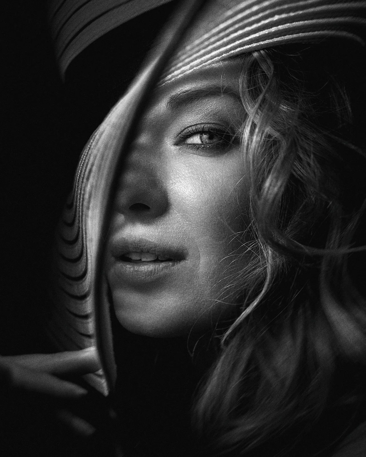 Close-up black and white portrait of a woman wearing a wide-brimmed hat, with curled hair partially covering her face, looking directly at the camera.