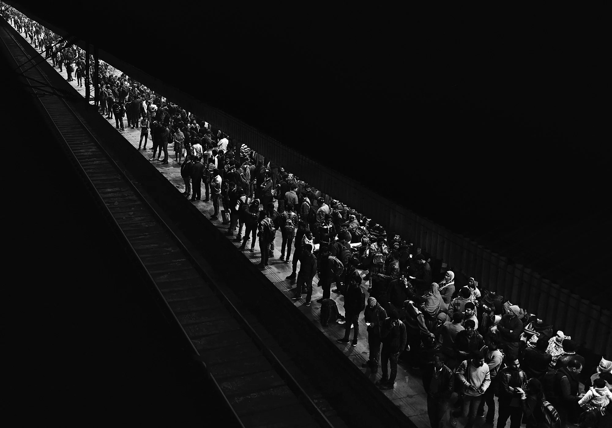 A long line of people waiting on a train platform at night, with most individuals standing and some sitting, all with backpacks, under a dark sky.