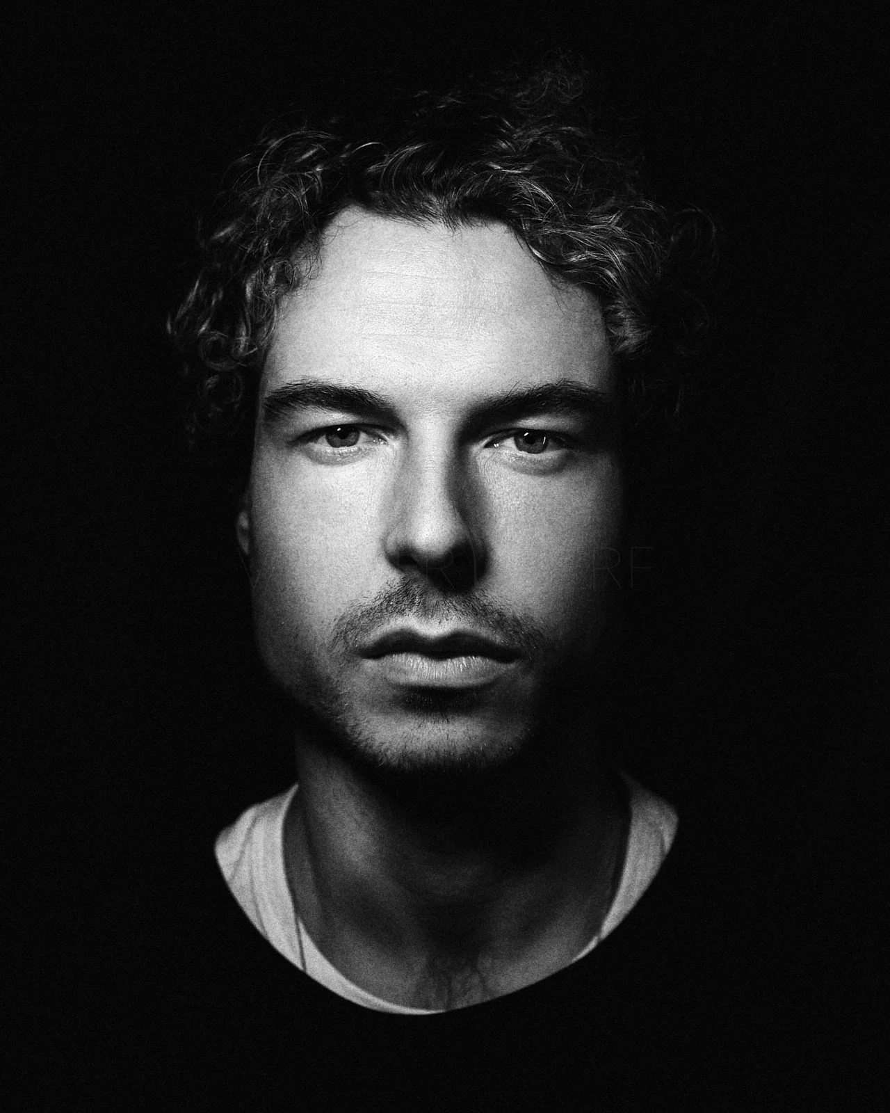 Black and white portrait of a young man with curly hair, intense gaze, and slightly parted lips, against a dark background.