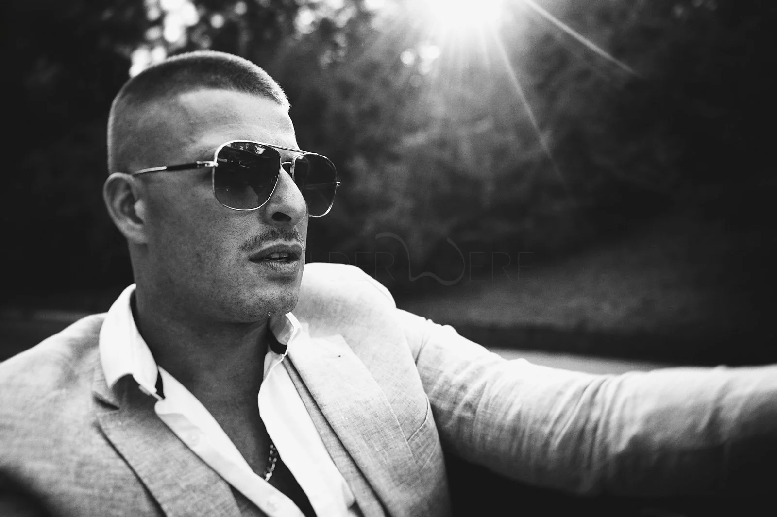 A man with short hair wearing dark sunglasses and a light-colored suit with an open-collared shirt, sitting outdoors with sunlight shining in the background.