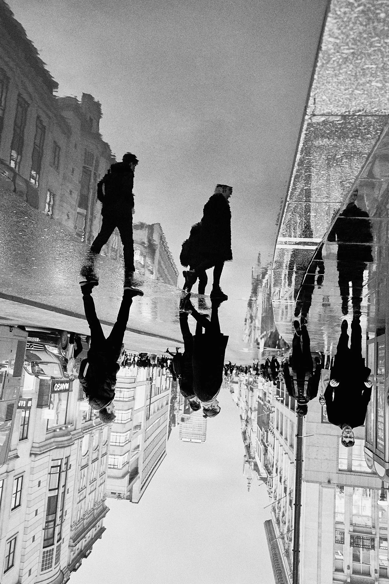 People walking on a city street with tall buildings in the background, reflected in a large puddle, black and white photo.