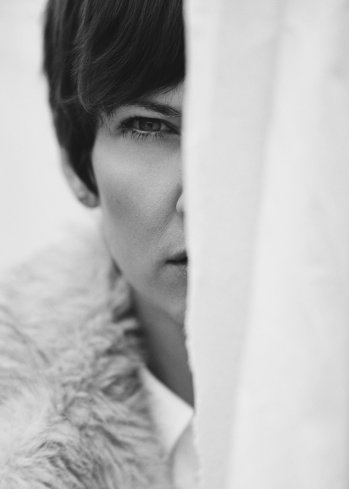 Black and white close-up photo of a person's face, mostly obscured behind a white curtain, with only one eye, part of their face, and a furry garment visible.