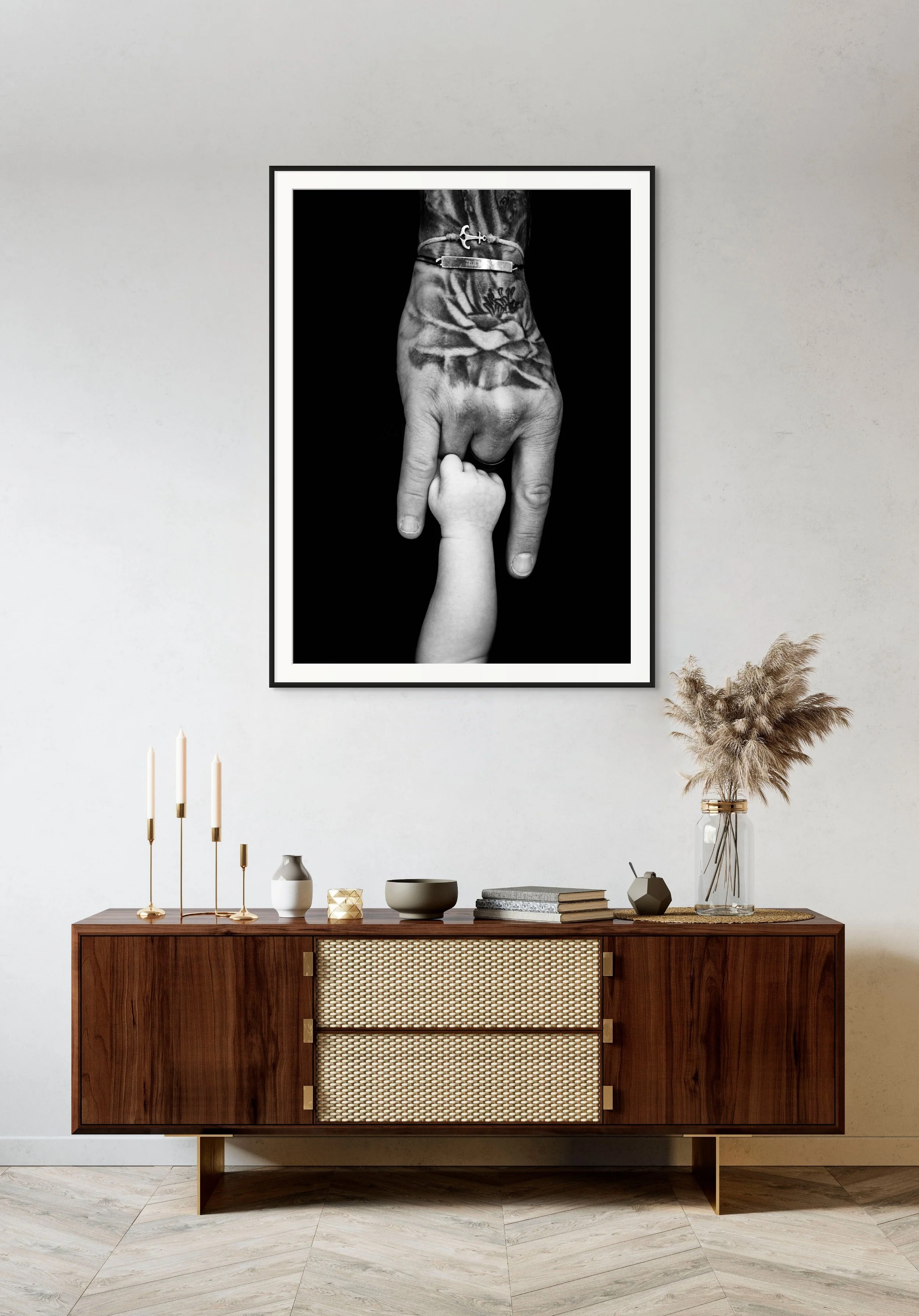 A black and white photo of a tattooed arm holding a child's hand, hanging on a wall in a modern living room with a wooden sideboard and decorative items.