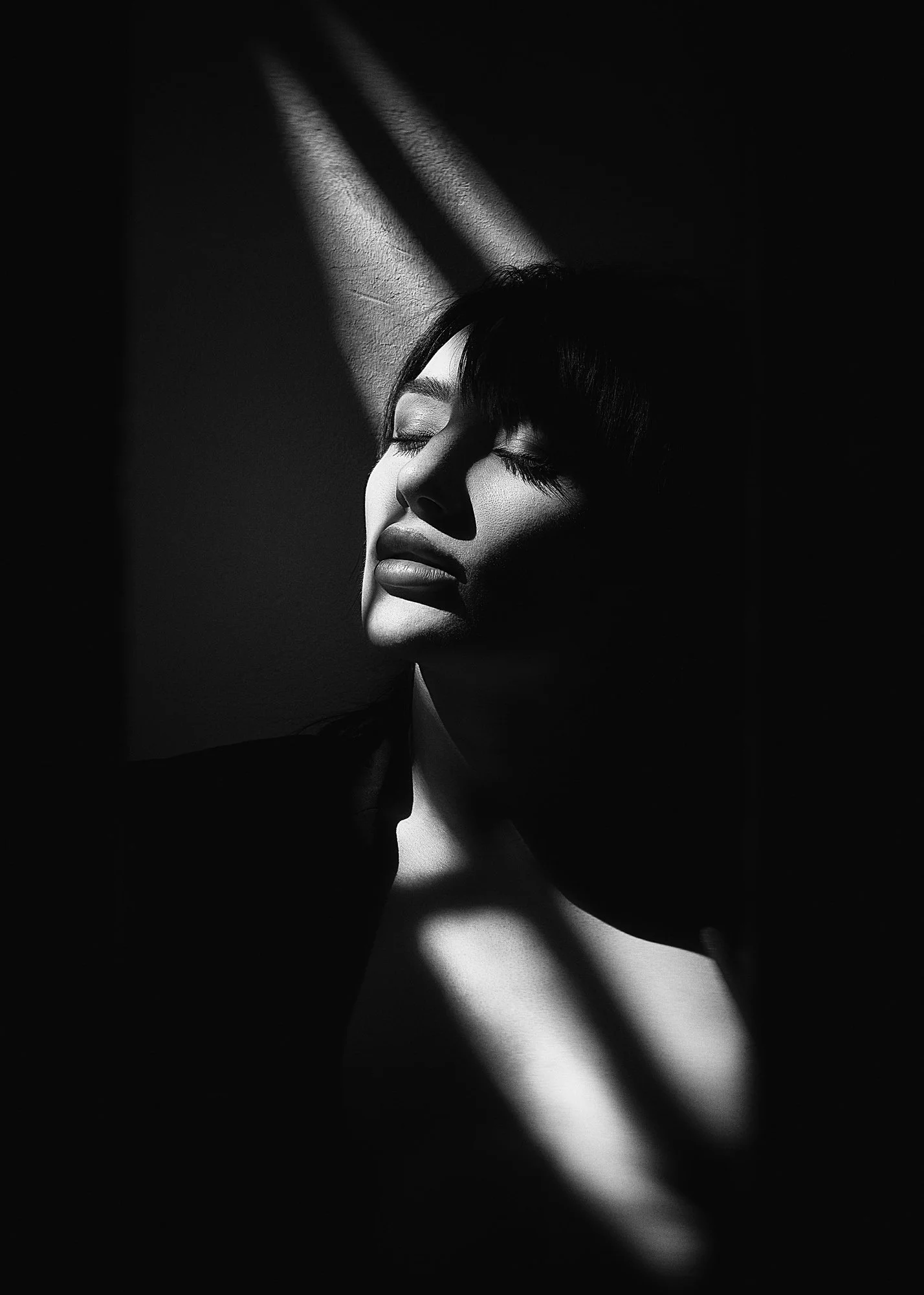 A black and white photo of a woman with eyes closed, illuminated by beams of light creating shadows on her face and neck.