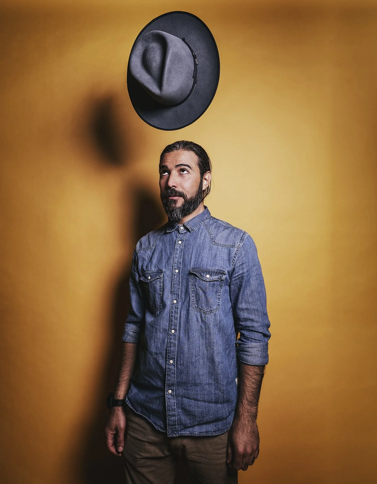 Man with a beard and long hair, wearing a denim shirt, looking up at a black hat with a gray fedora on top, hanging on a yellow wall.