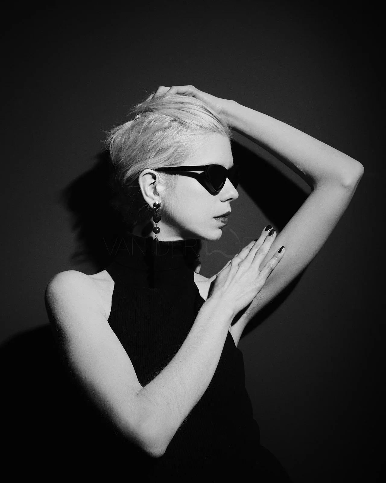 Black and white portrait of a woman with short blonde hair wearing dark sunglasses, a sleeveless black top, and earrings, with one arm raised and hand on her head, in a dramatic pose.