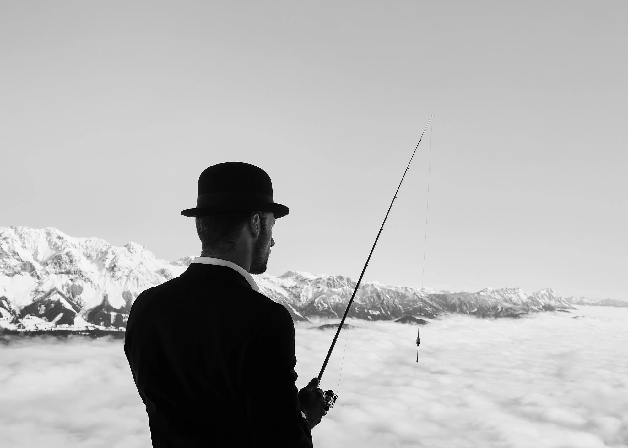 A man in a suit and bowler hat fishing above a sea of clouds with snow-covered mountains in the background.