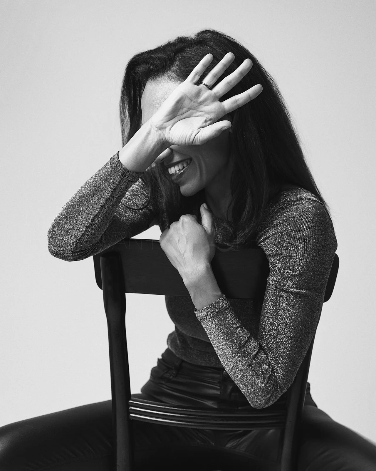 A woman with long dark hair smiling, sitting on a chair, covering part of her face with her hand, in a black and white photograph.