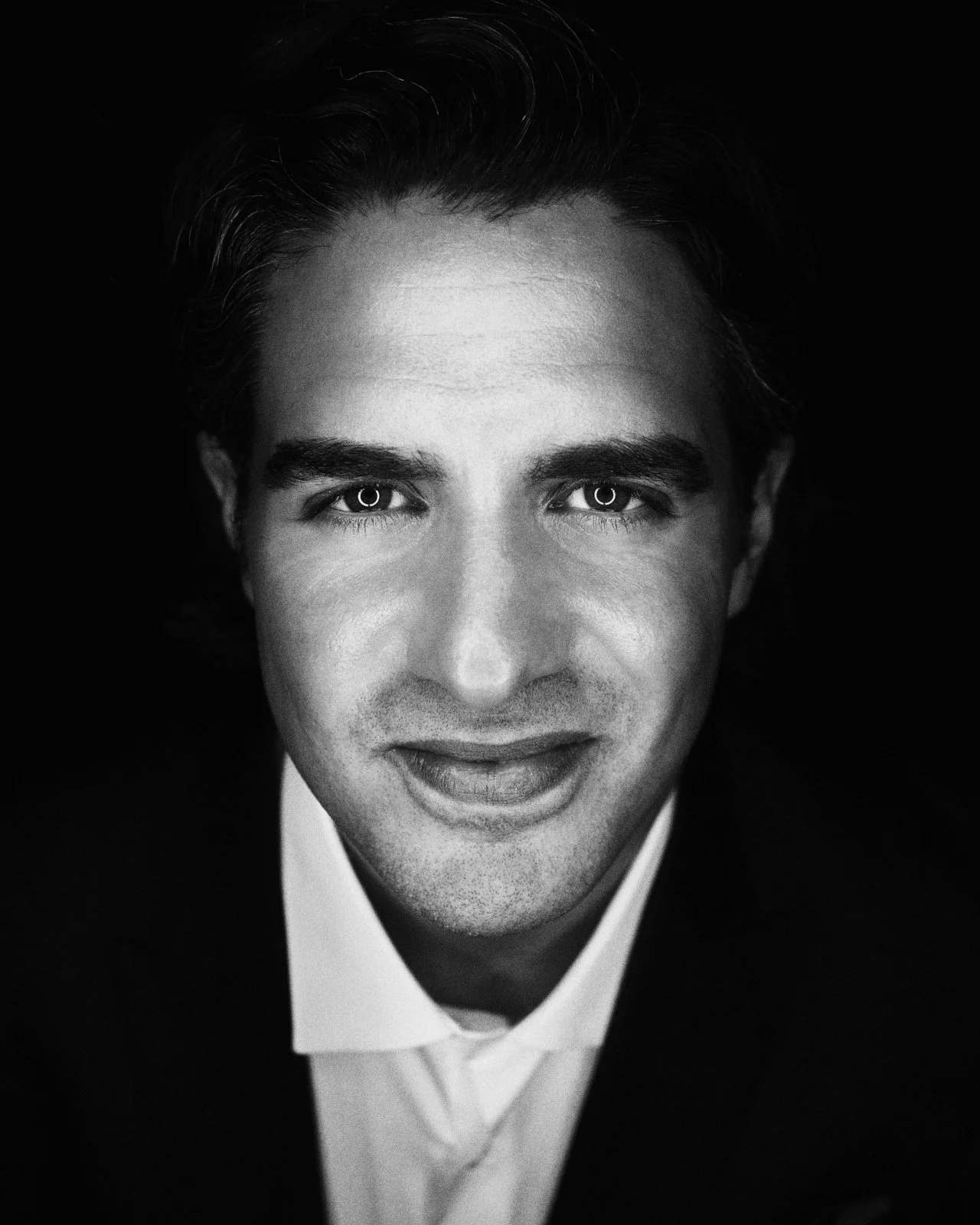 Black and white close-up portrait of a man with dark, slicked-back hair, wearing a white shirt and black jacket, smiling slightly against a black background.