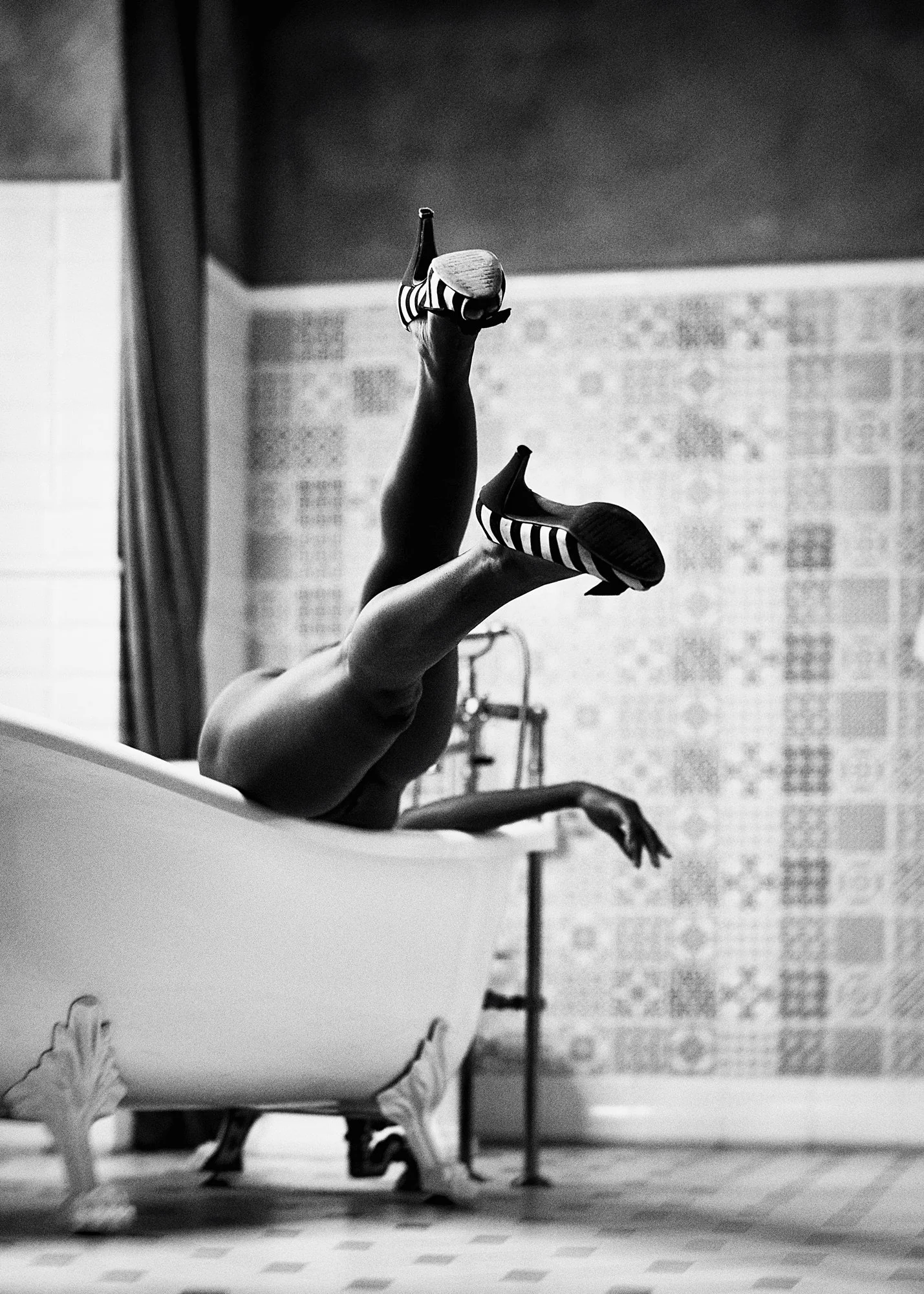 A person falls into a clawfoot bathtub, wearing striped high heels, with their legs and feet in the air and arms outstretched, in a room with patterned wallpaper.
