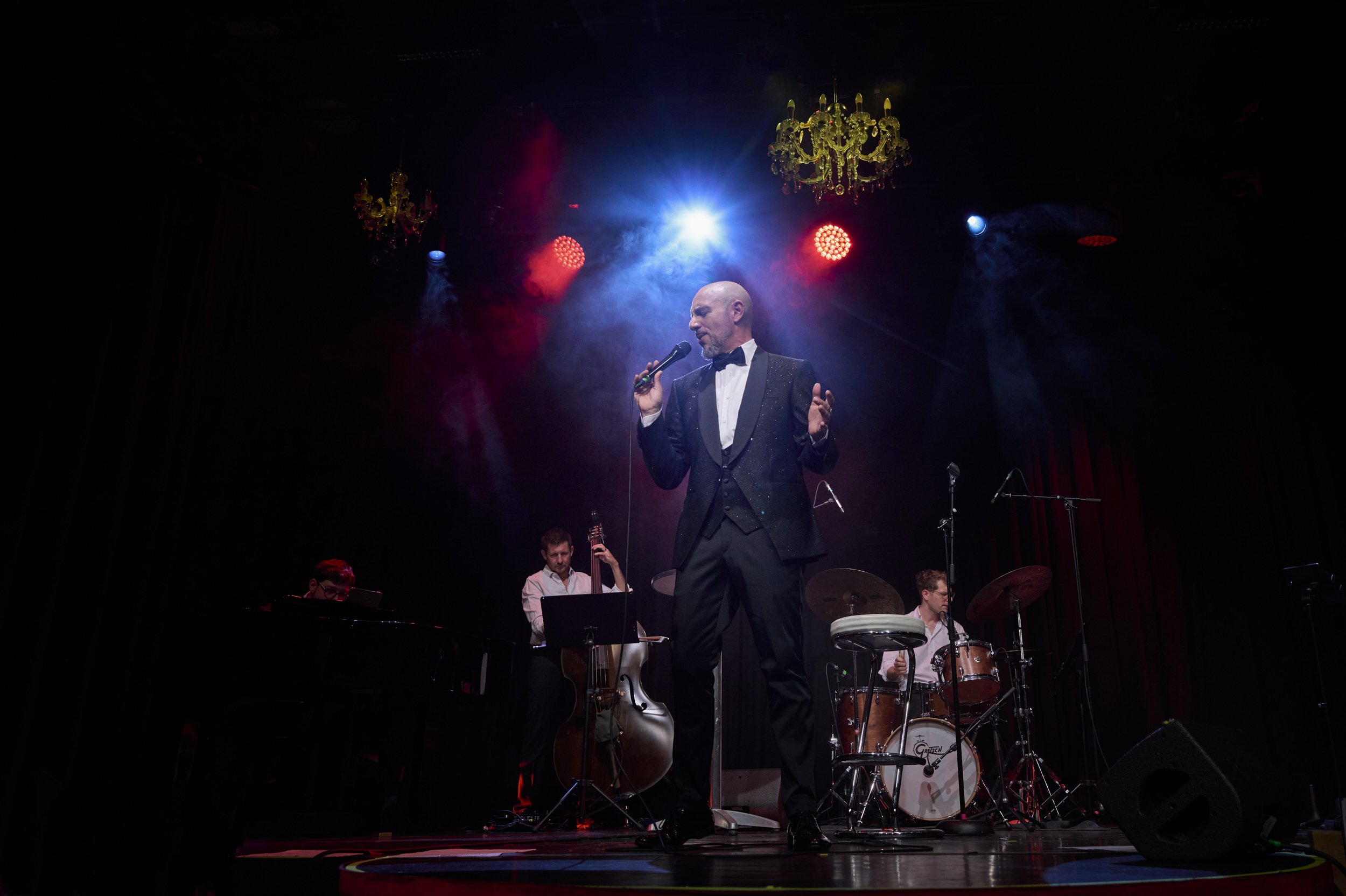 A man in a tuxedo singing into a microphone on stage with a band playing instruments behind him, colorful stage lighting, and chandeliers hanging from the ceiling.