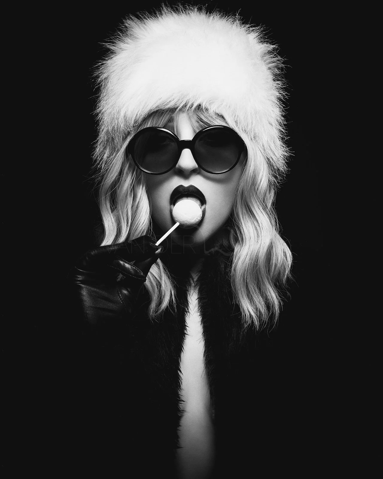 Black and white photo of a woman wearing a large fuzzy hat, dark sunglasses, and gloves, holding a lollipop near her mouth.