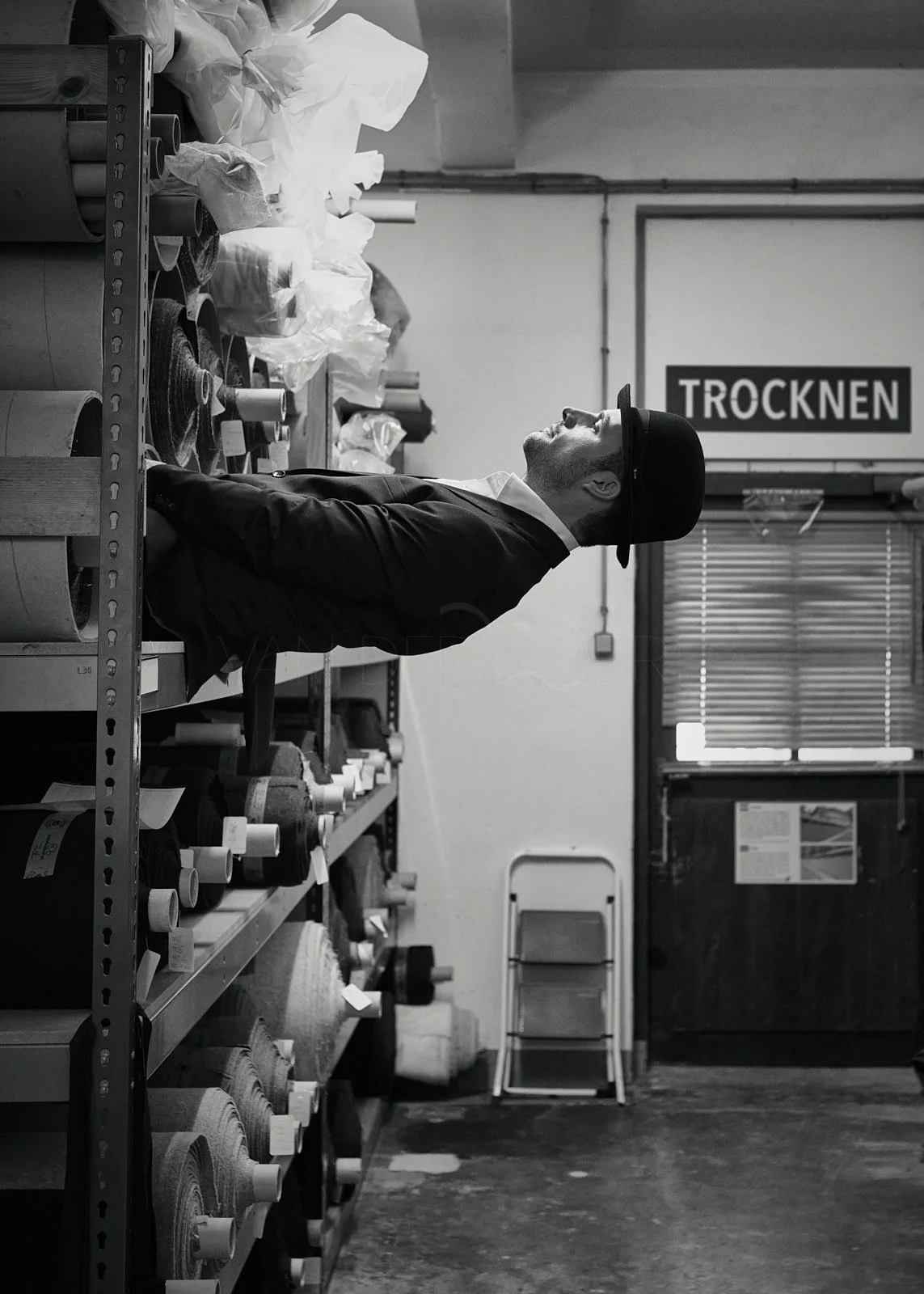 A man in a hat and coat standing in a store aisle filled with rolls of fabric or paper, looking upward. The store has a sign that says 'TROCKNEN' in the background.
