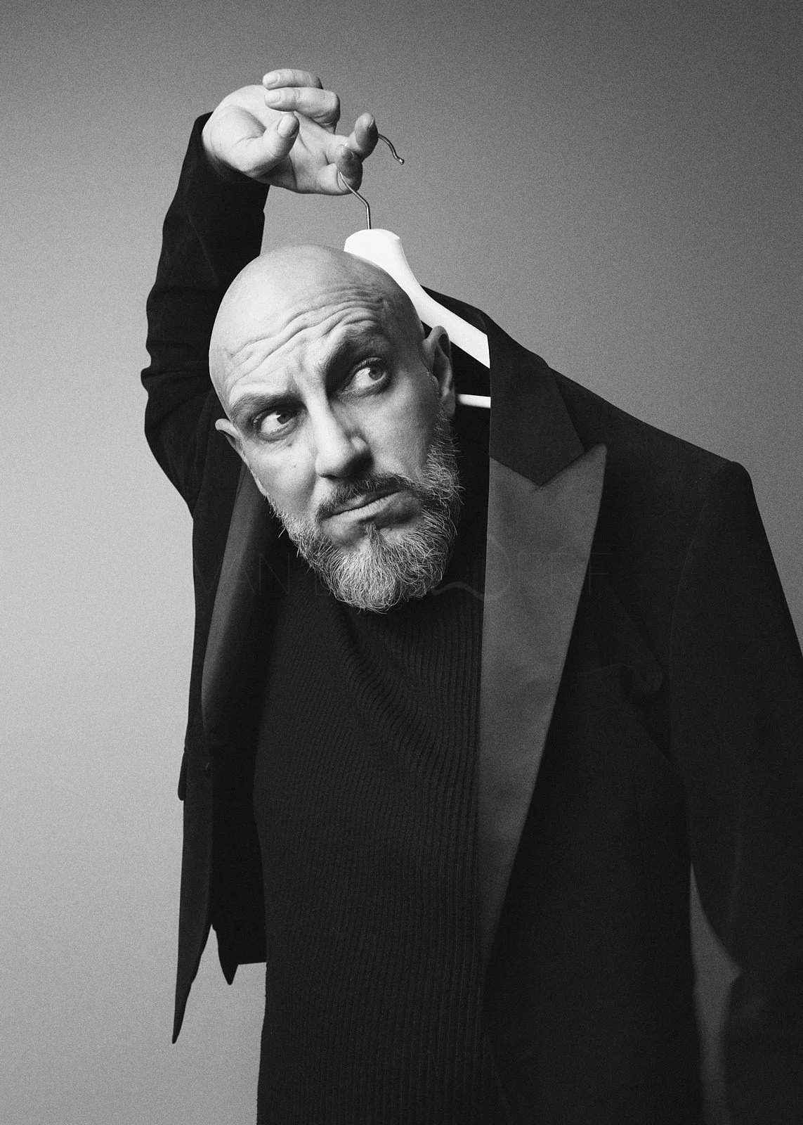 A black-and-white photograph of a bald man with a beard, wearing a tuxedo, looking to the side while holding a clothing hanger near his head.