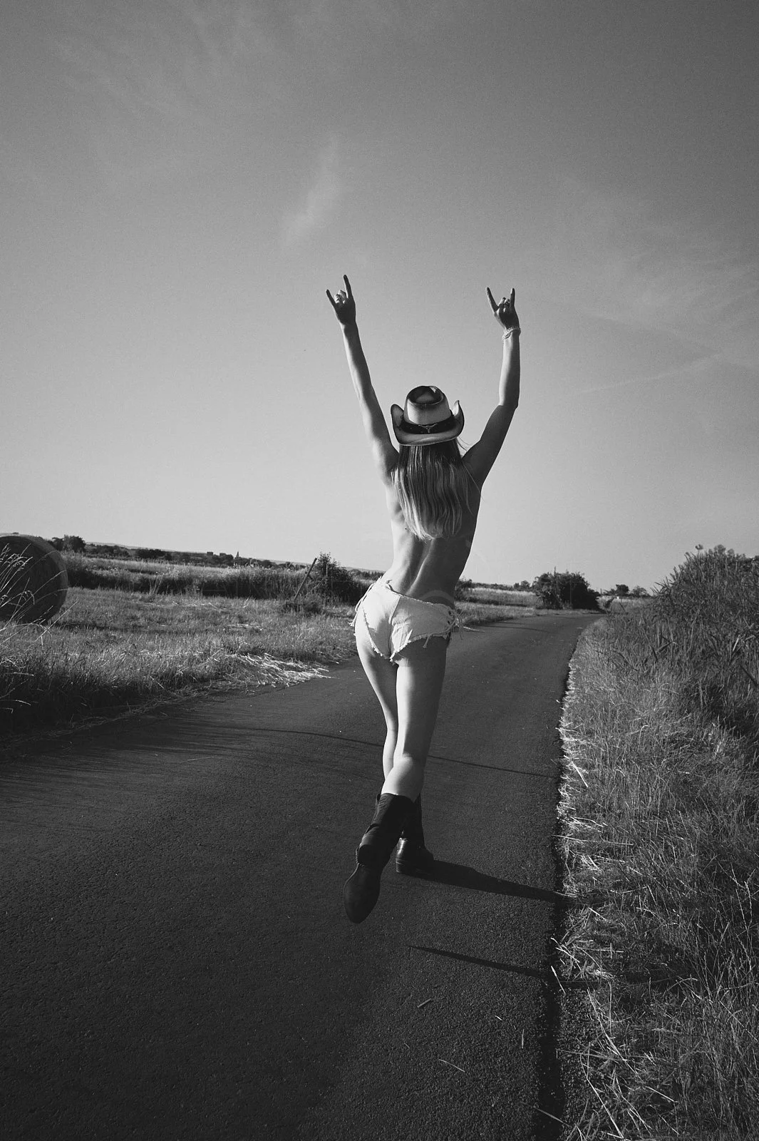 A woman with long hair wearing a cowboy hat, shorts, and boots, jumping in the air on a rural road with grassy fields on either side, waving her arms with peace signs in a black and white photo.