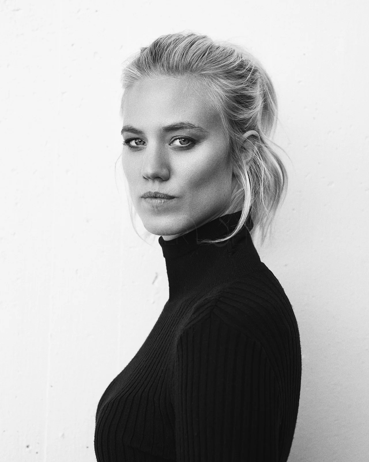 A black-and-white portrait of a woman with blonde hair tied back, wearing a black turtleneck sweater, looking directly at the camera against a plain white background.
