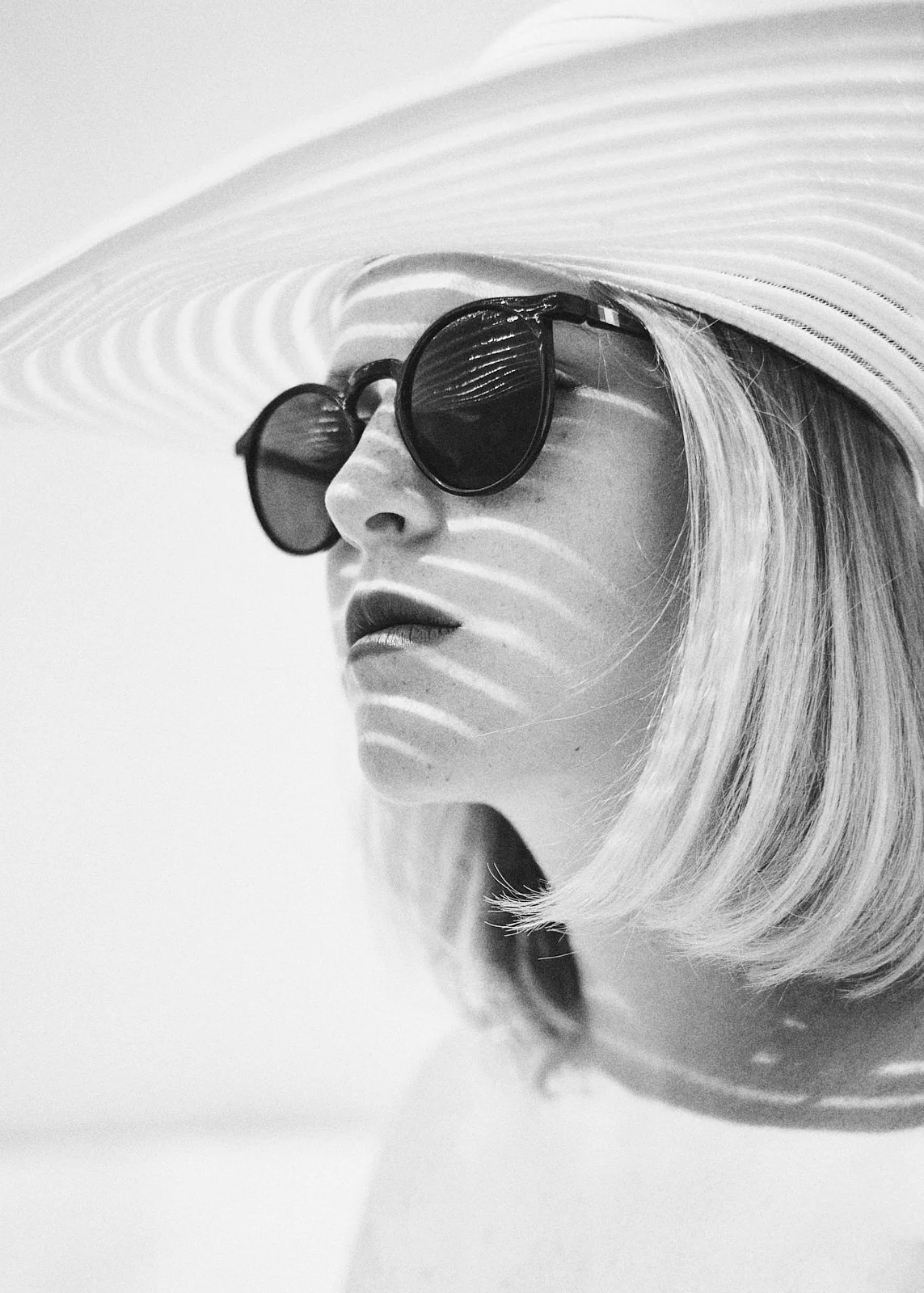 A woman wearing large round sunglasses and a wide-brimmed hat, with striped shadows falling across her face, in black and white.