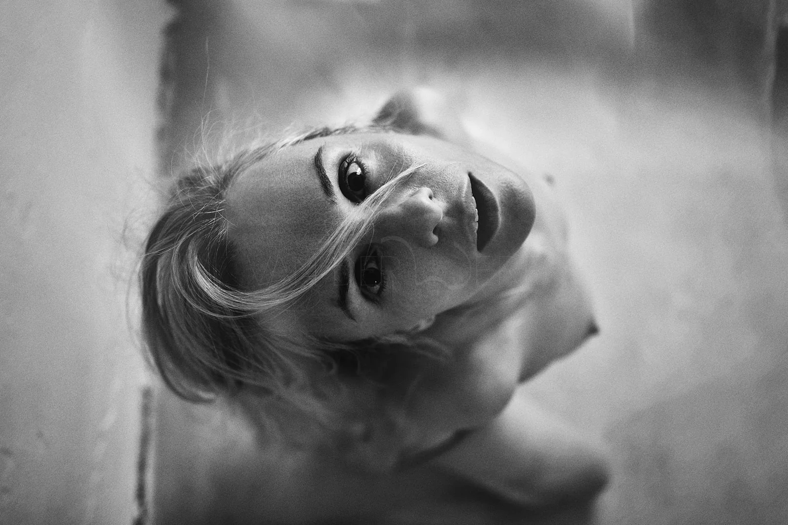 Black and white photo of a woman with blonde hair lying on the ground, looking directly at the camera with an intense gaze.