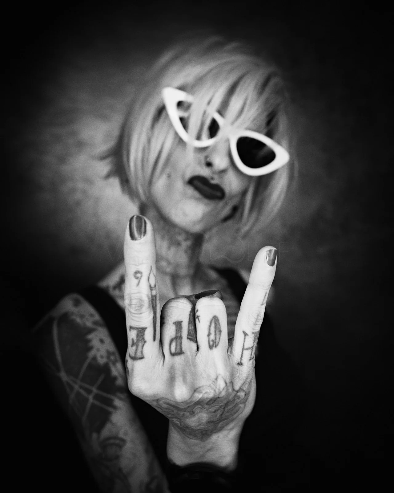 Black and white photo of a woman with short blonde hair wearing white cat-eye sunglasses and dark lipstick, showing her middle finger with tattoos, and giving a rebellious gesture.