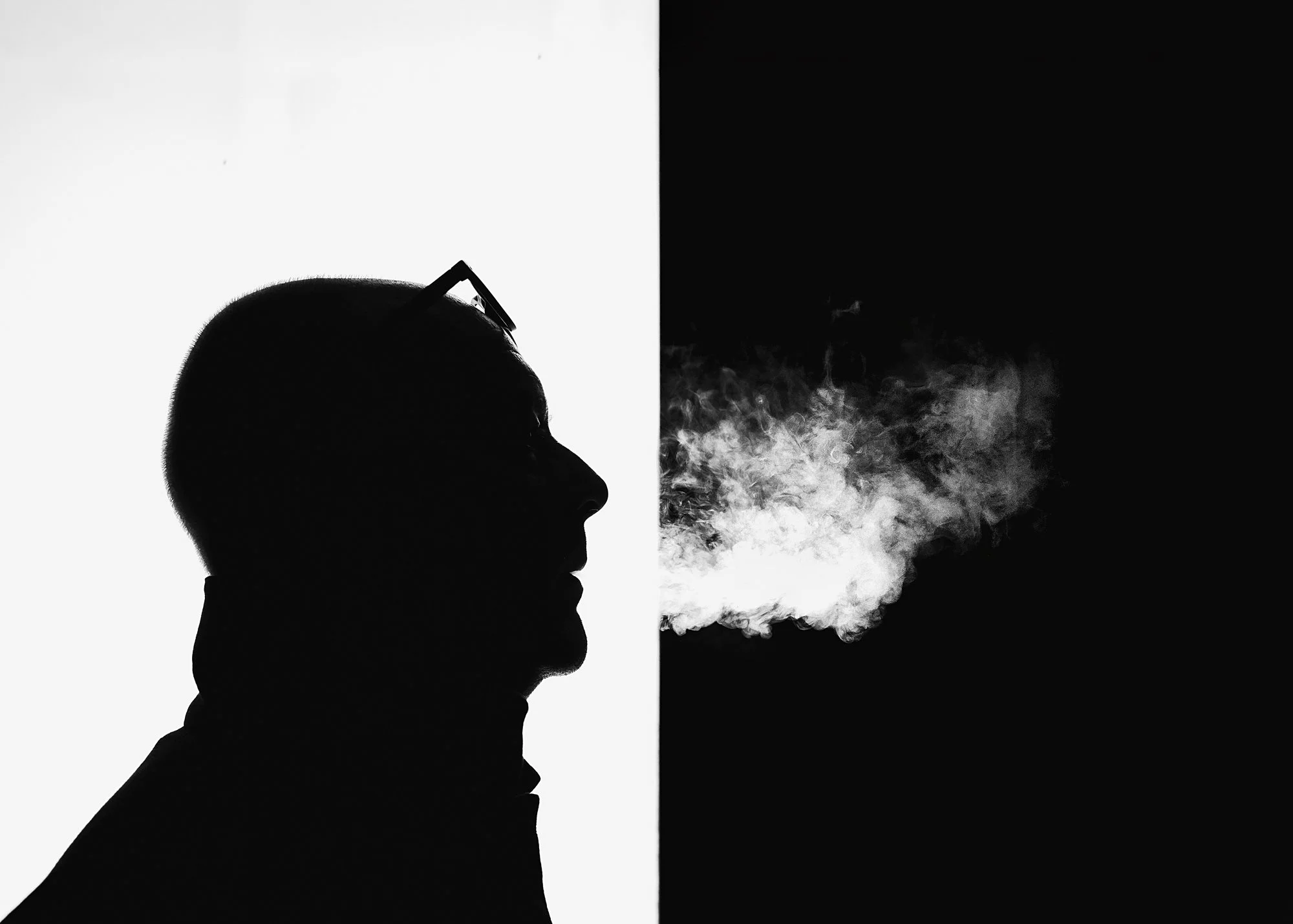 Silhouette of a man wearing glasses, with smoke or vapor coming from his mouth, against a background split into white and black.