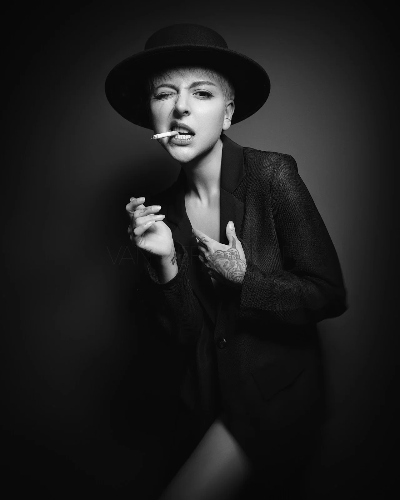 Black and white portrait of a woman wearing a hat, with short hair, holding a cigarette in her mouth, winking, and pointing to herself, against a dark background.