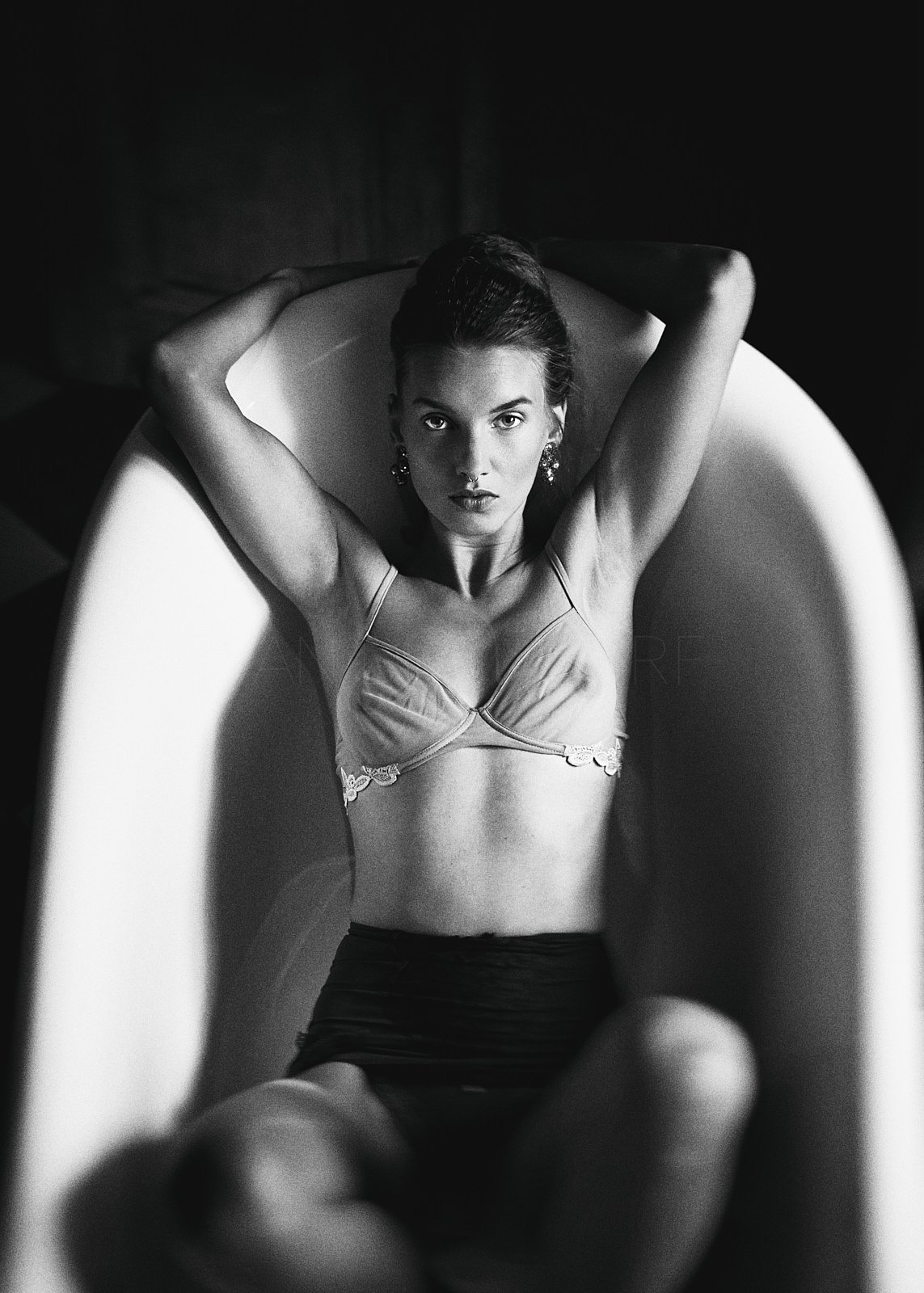 Black and white photograph of a woman lying back in a chair with her arms resting behind her head, wearing a bra and skirt, looking directly at the camera.
