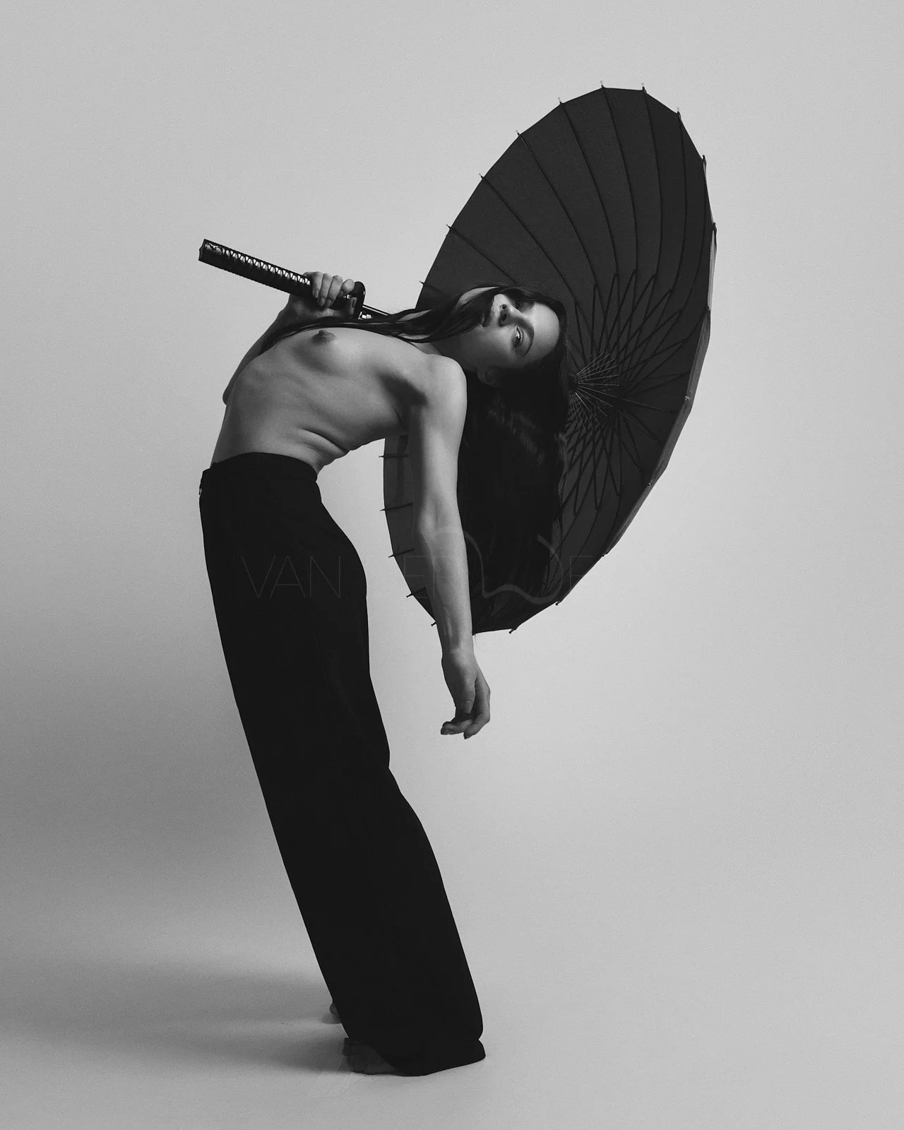 A black and white photo of a topless woman holding an umbrella, leaning backward with long dark hair, wearing loose black pants, against a plain background.