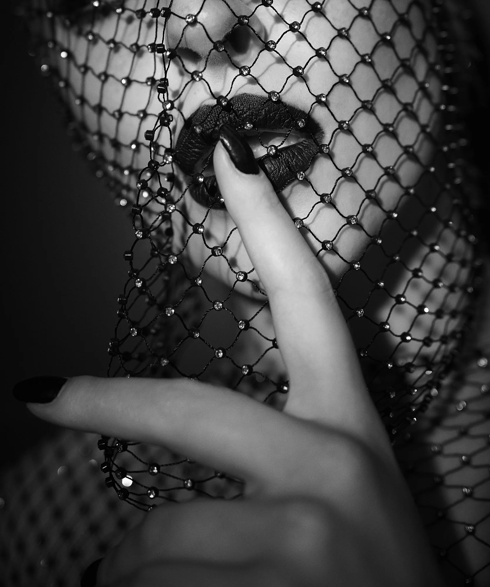 A black and white close-up photo of a woman's face covered with a black fishnet veil. She is biting her finger and has dark lipstick and nail polish.