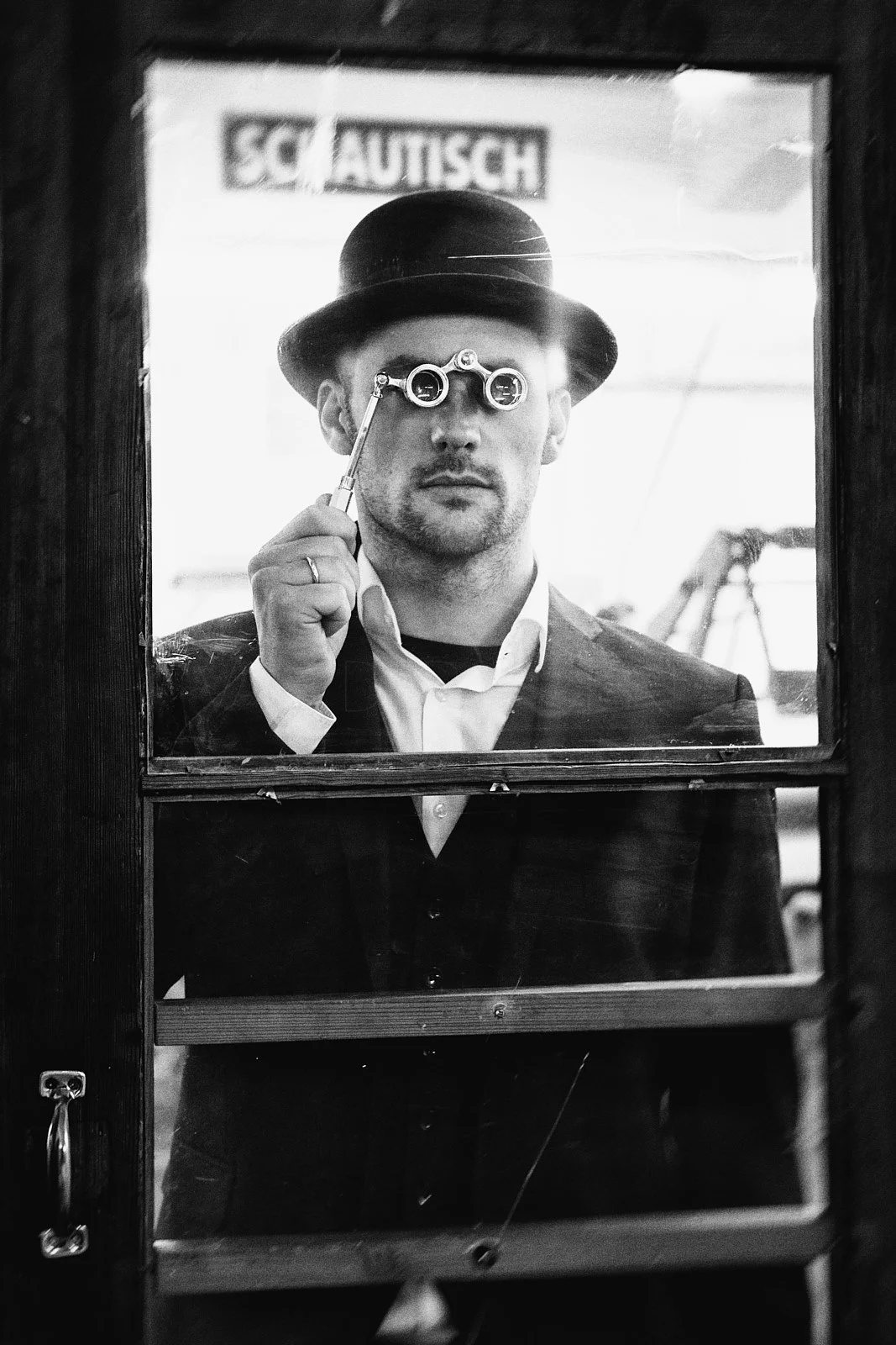 Black and white photo of a man seen through a door window, wearing a bowler hat and suit, examining with a monocle and holding a small tool, with a blurred sign in the background.