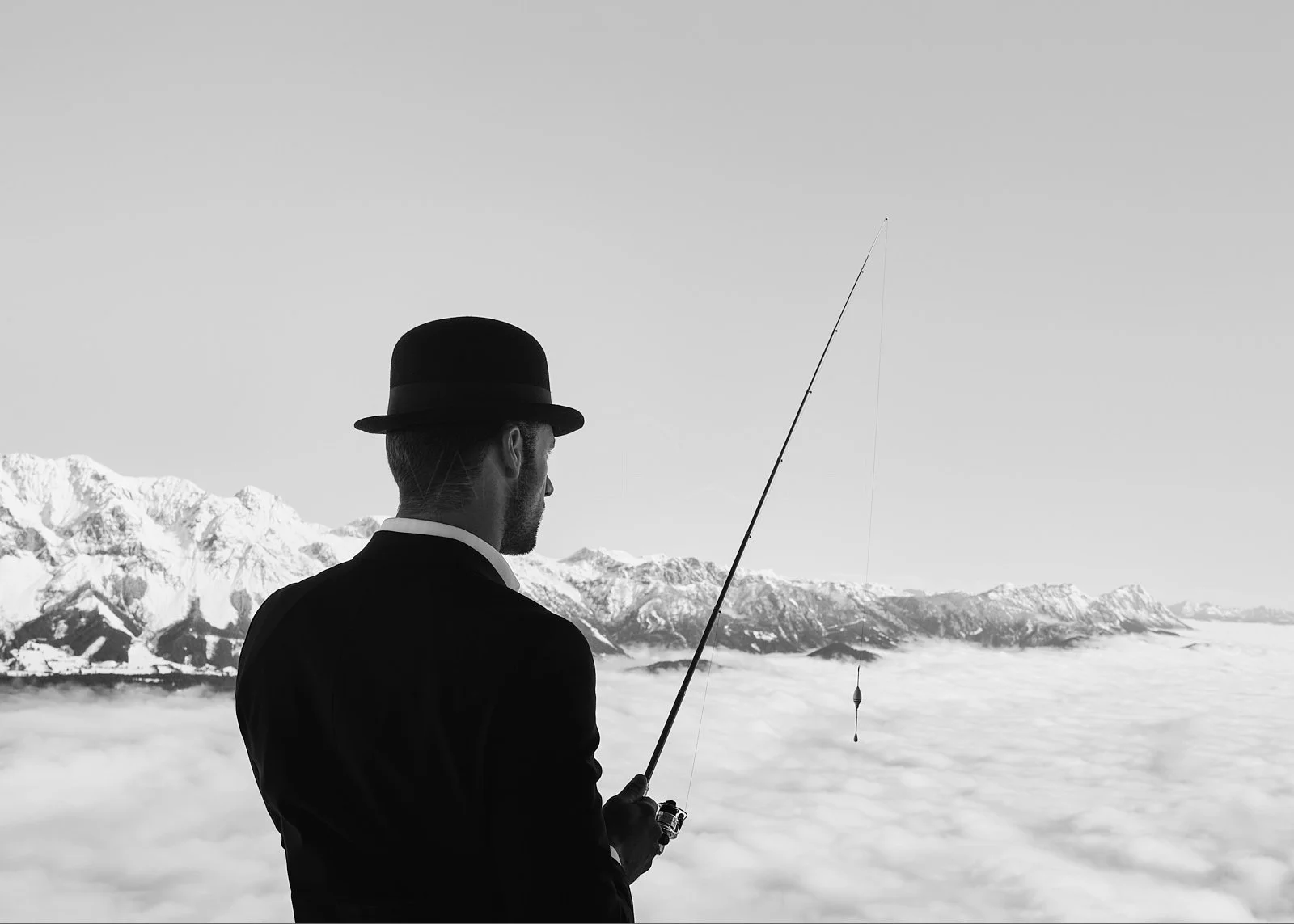 A man wearing a bowler hat and suit, holding a fishing rod, stands among clouds with a mountain range in the background.