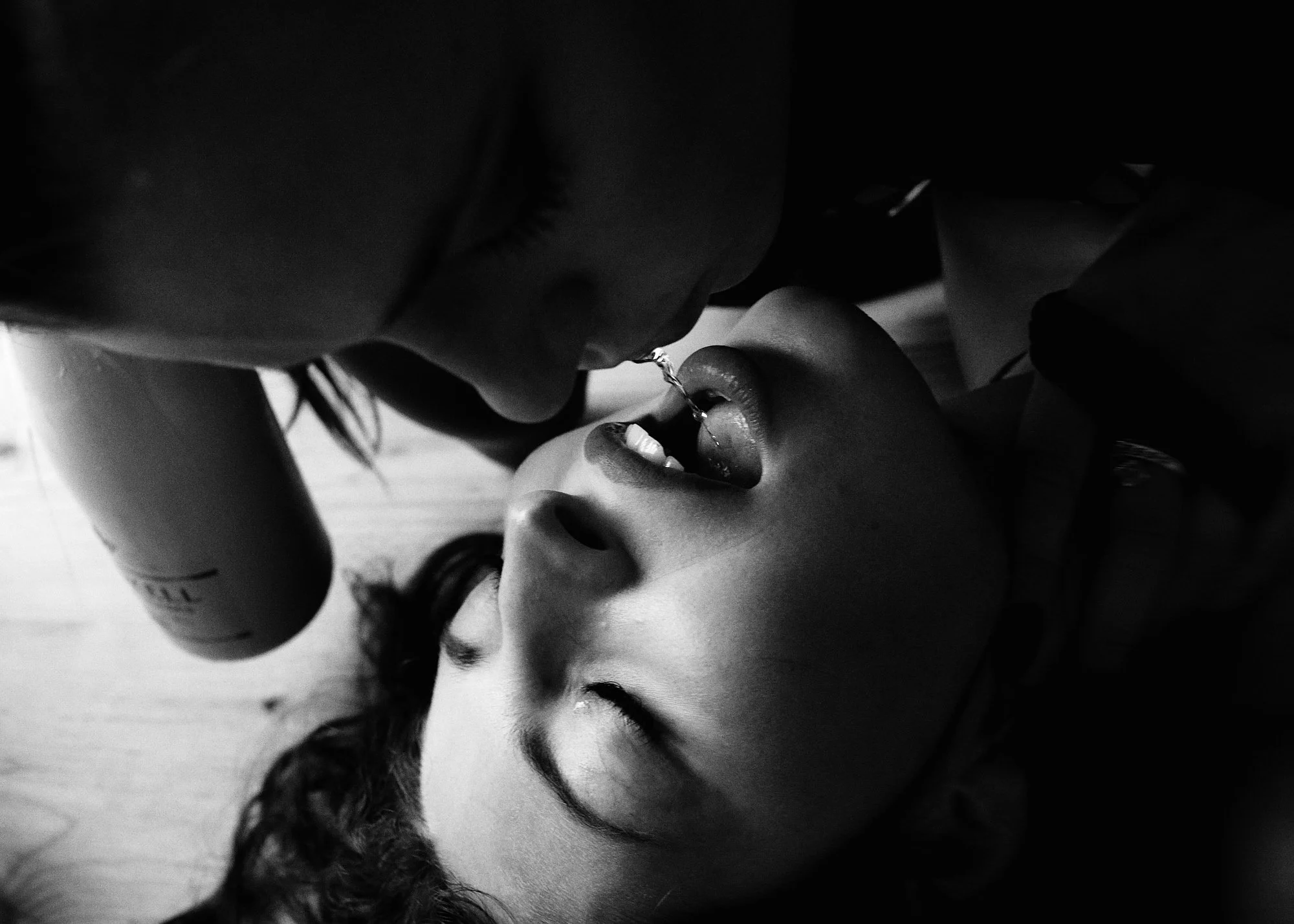 Black and white image of one person dripping liquid from their mouth into another person's mouth, both lying on a surface.