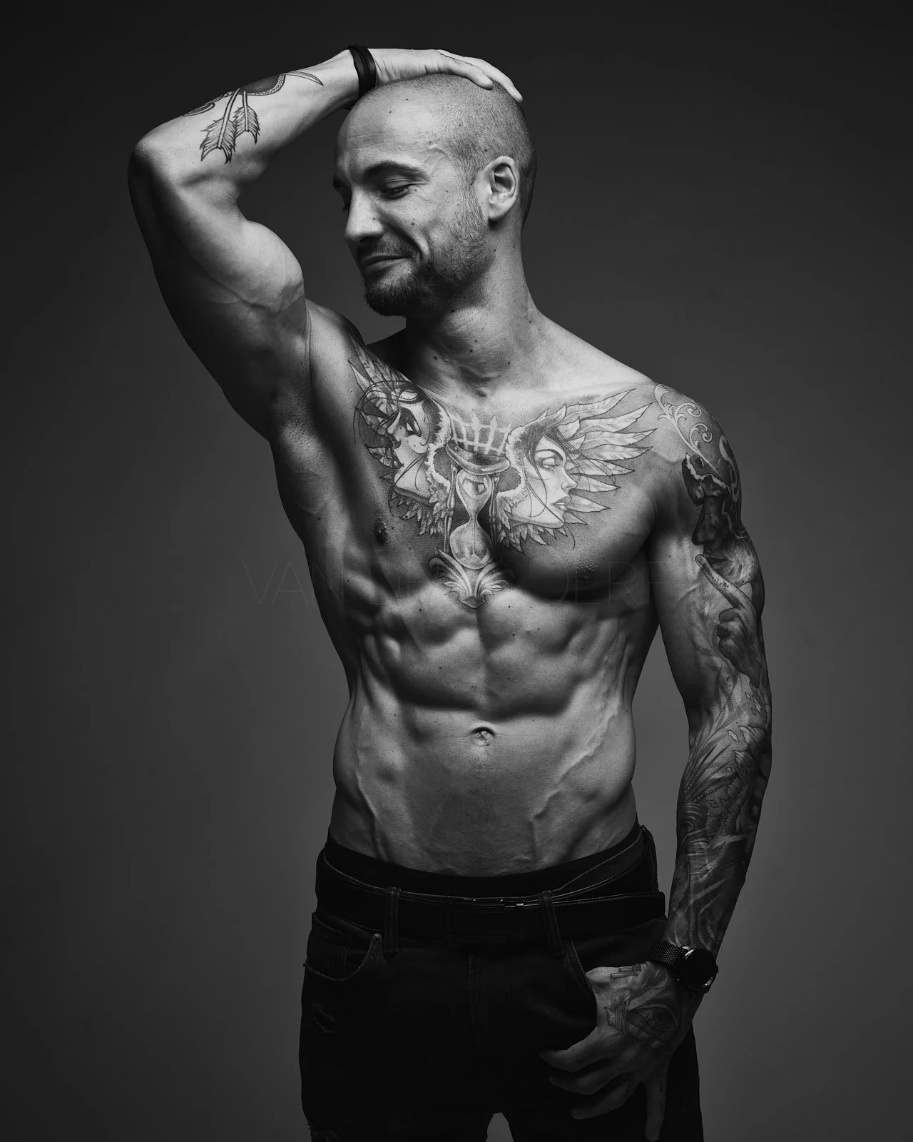A shirtless man with tattoos posing with one arm behind his head, smiling softly.