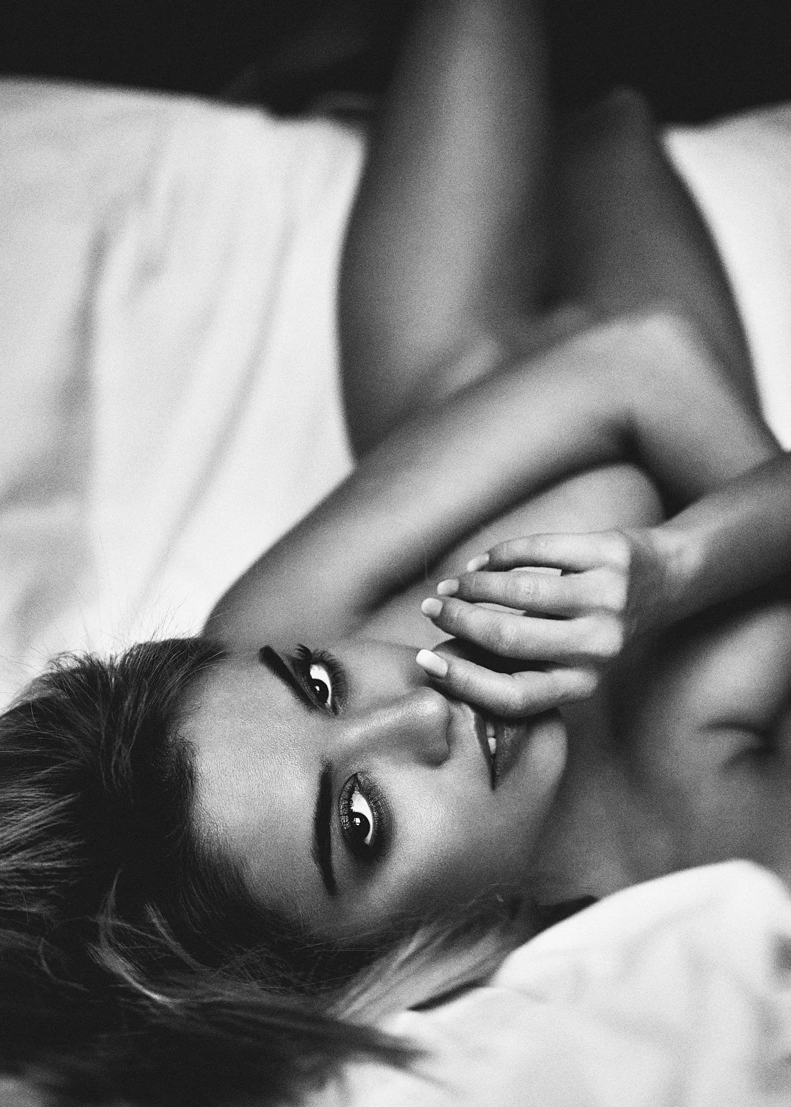A woman lying on her back on a bed, with one hand near her face, looking directly at the camera. The photo is in black and white.